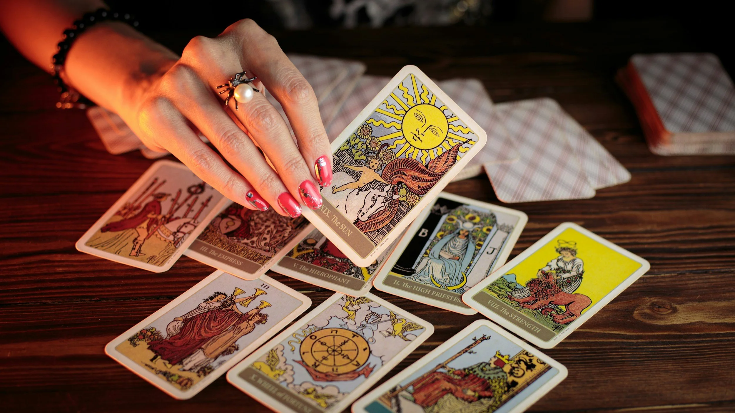 A person with painted nails and jewelry holds a tarot card titled 'The Sun' facing upward, surrounded by other tarot cards spread on a wooden surface, with some face-down cards nearby.
