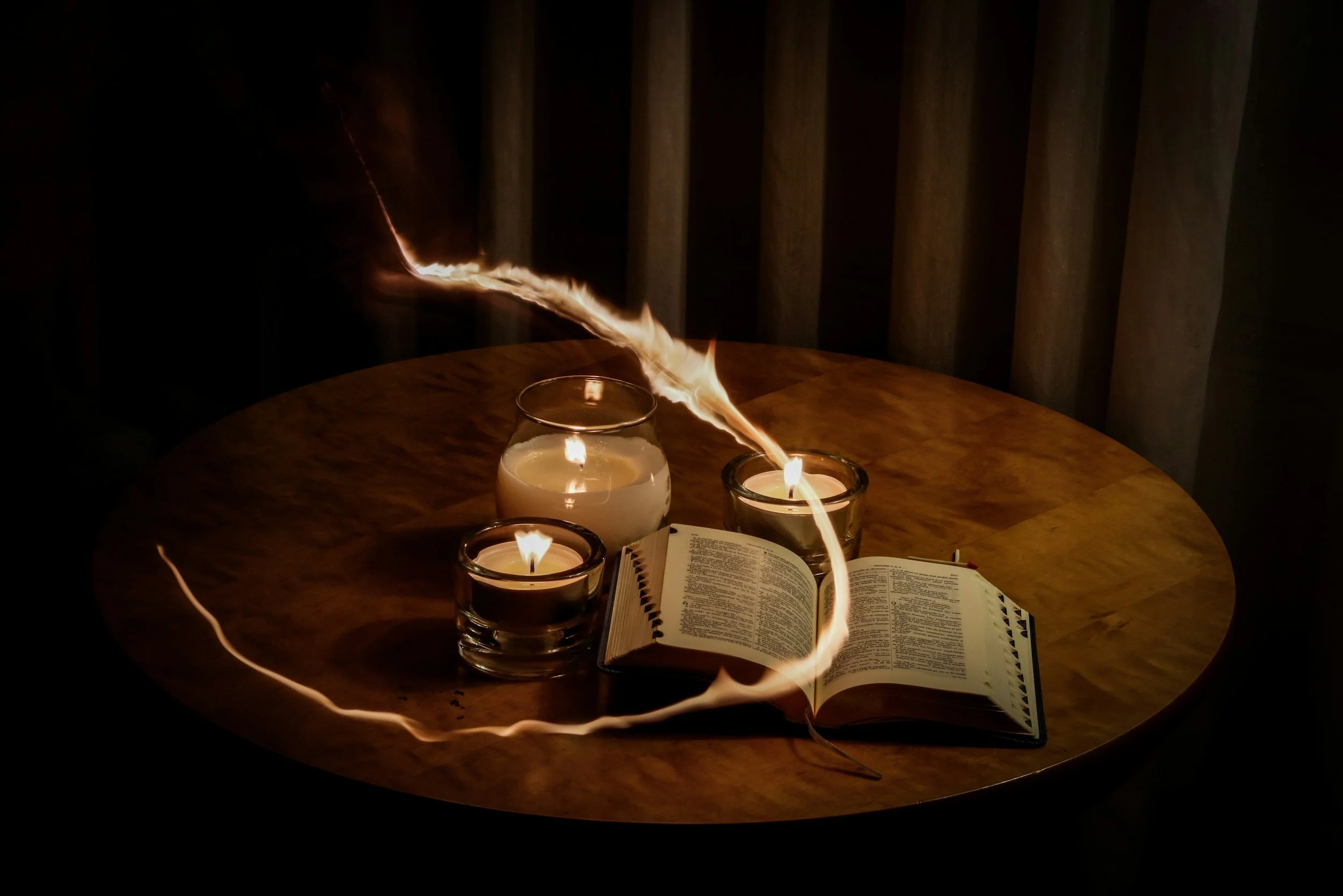 A round wooden table holding an open book, two lit candles in glass holders, a glass of water with a candle inside, and a lightning bolt-shaped streak of light passing over the objects. Medium psychic readings for all online.