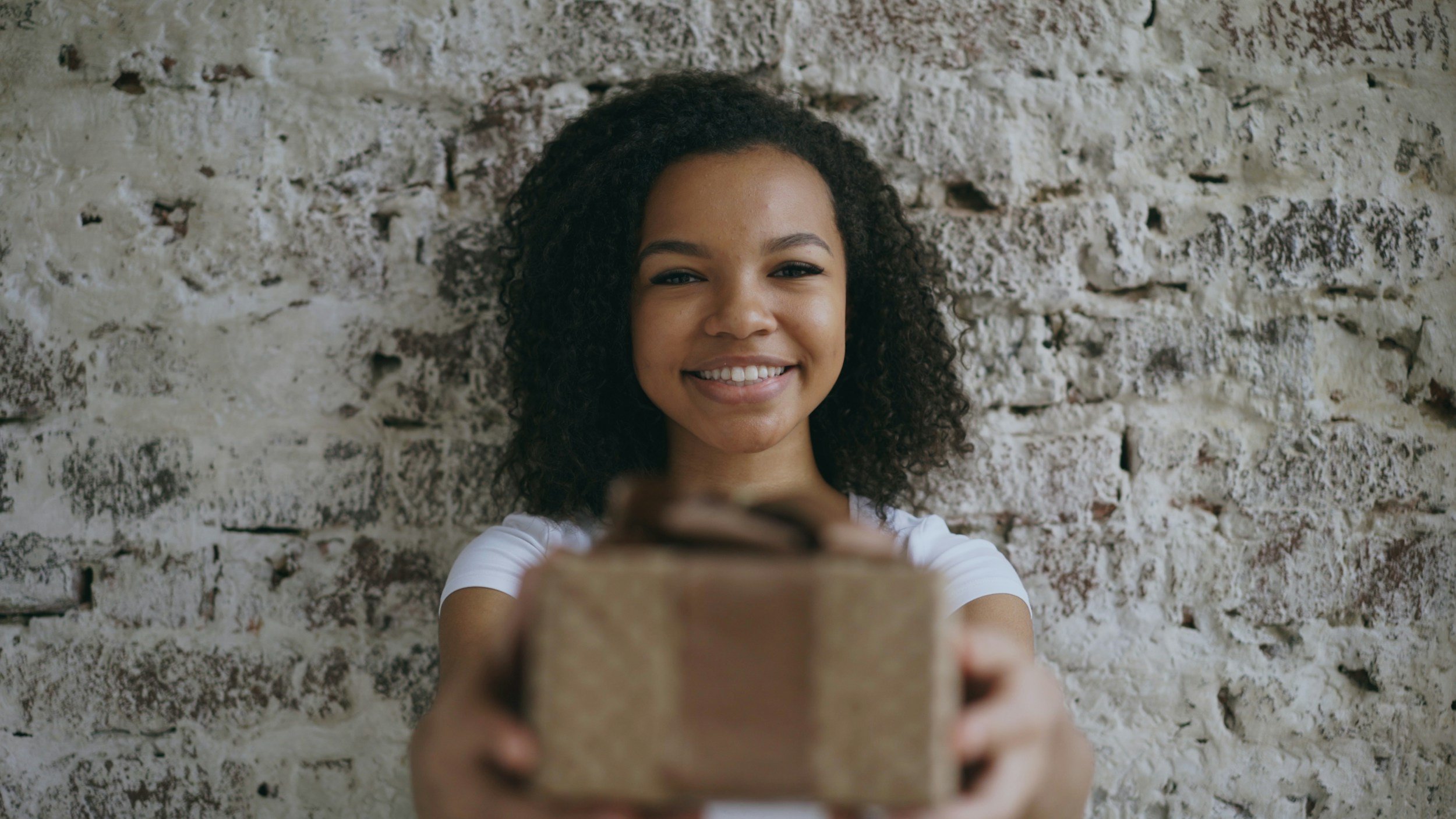 A smiling young woman with curly hair taking a selfie against a textured brick wall.