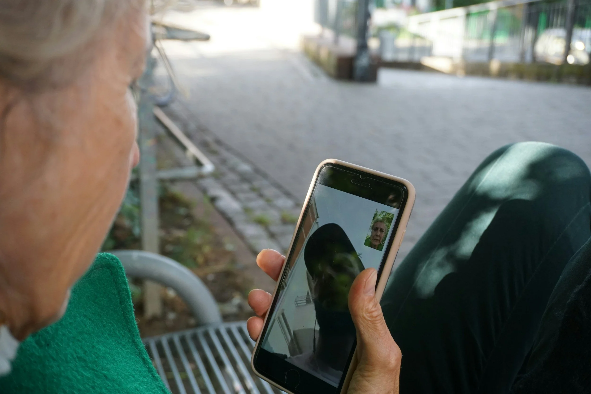 An elderly person sitting outdoors on a park bench, looking at a smartphone, which displays a video call with another person. The scene shows a pathway with a railing and some greenery in the background.