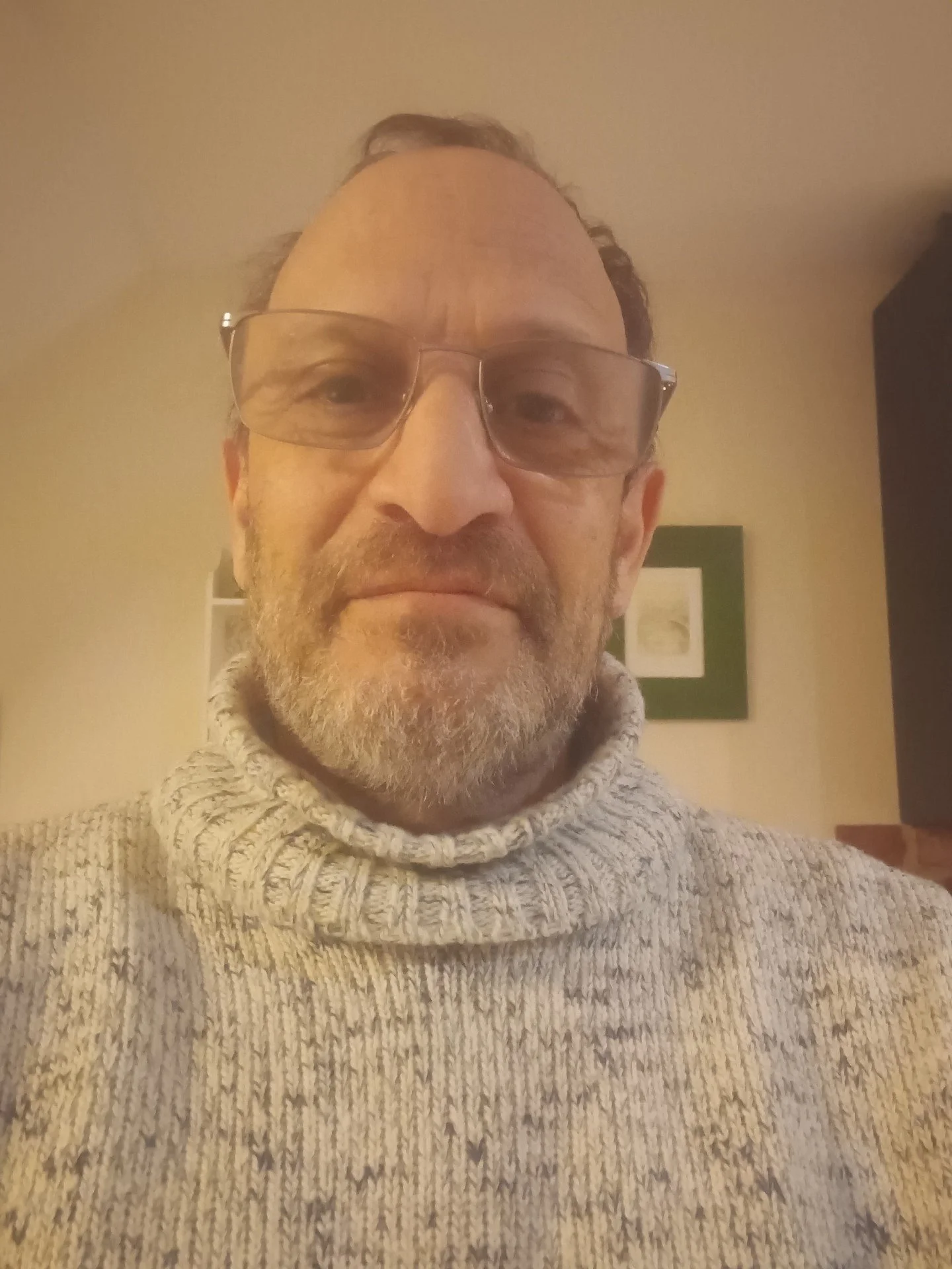A middle-aged man with glasses, a beard, and grey hair wears a thick, light-colored knit turtleneck sweater, with a neutral expression, standing indoors.