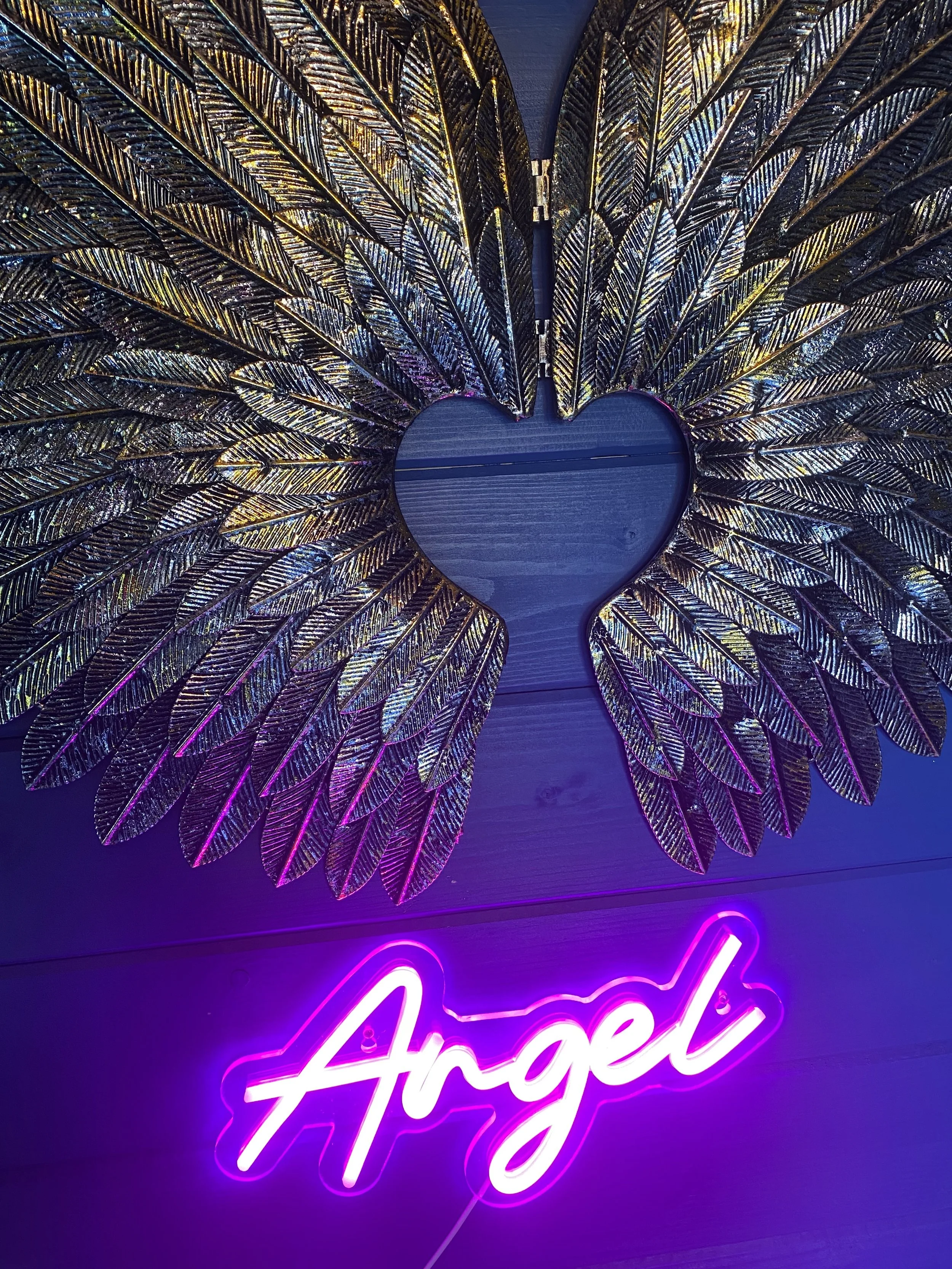 Decorative wall with gold feather wings forming a heart shape and a neon pink sign that says 'Angel' underneath.