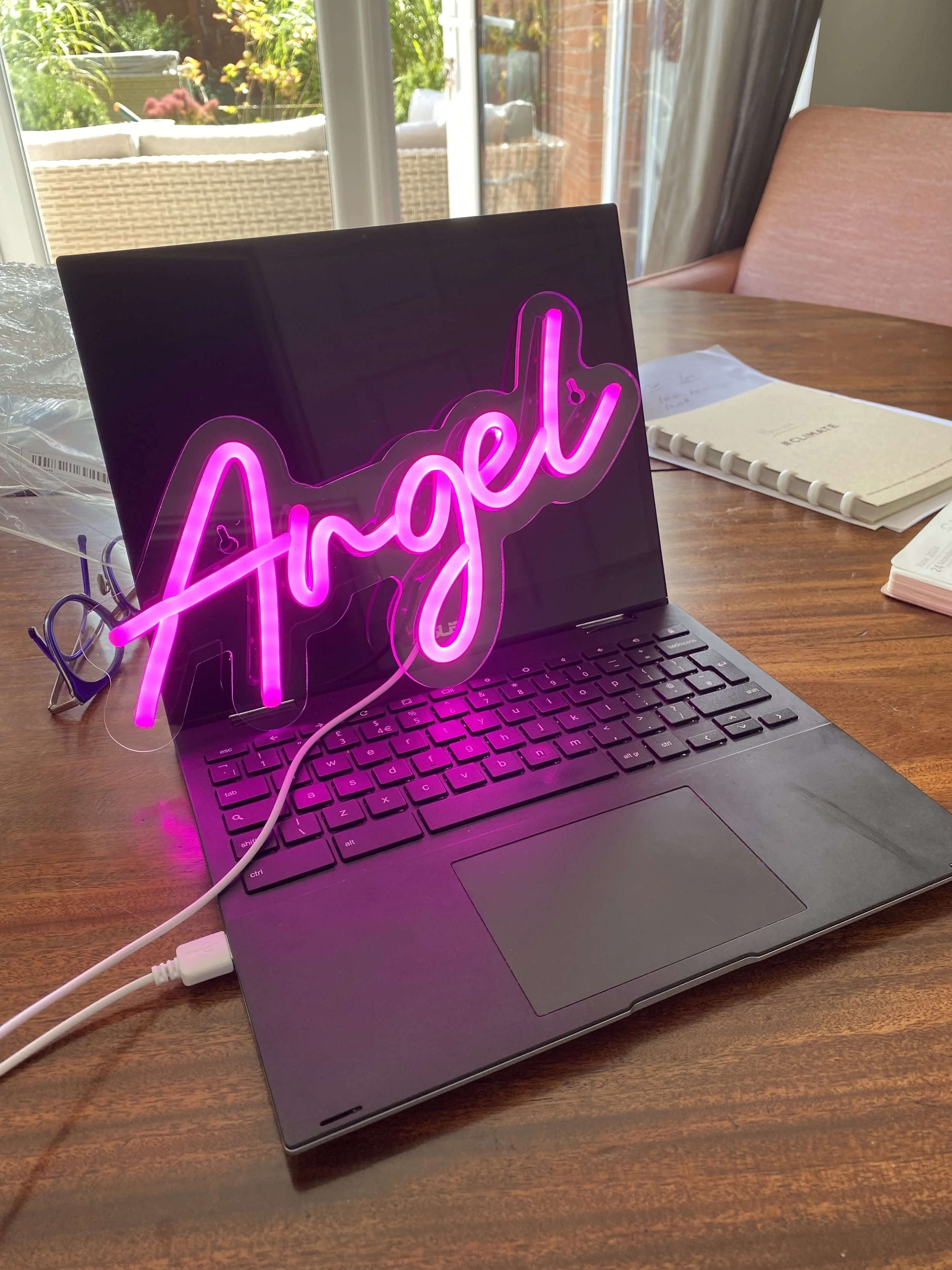A laptop on a wooden table with a pink neon sign that says 'Angel' leaning against the screen. On the table, there are also some notebooks, papers, and a pair of black-rimmed glasses. The background shows a bright outdoor patio with chairs and greenery visible through large windows.