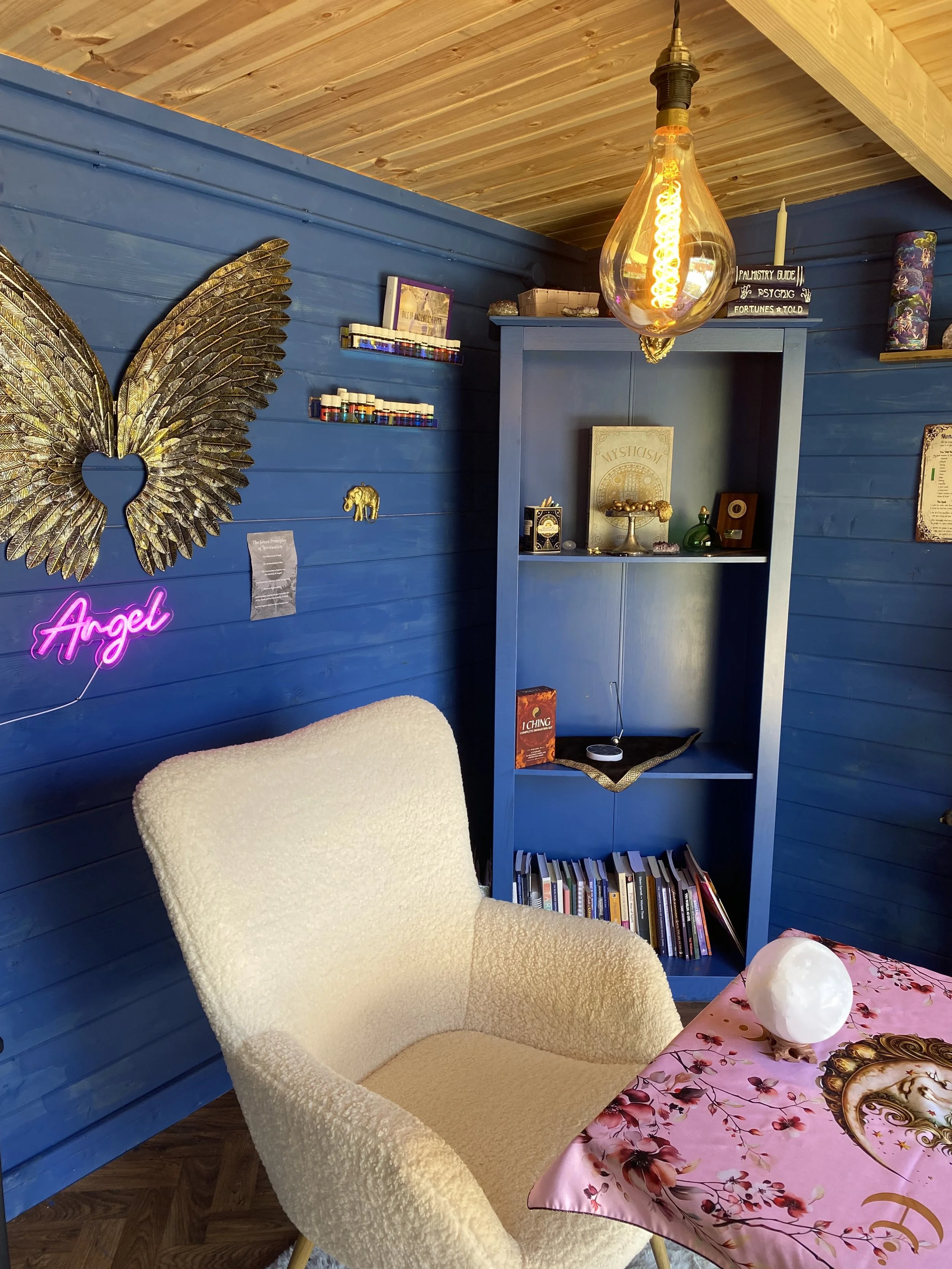 A cozy room with blue wooden walls and a wooden ceiling, decorated with eclectic items. There is a white, fluffy armchair in front of a pink tablecloth-covered table with a decorative orb. On the wall, a neon pink sign reads 'Angel' and a large gold butterfly wing art piece. A small bookshelf contains books, and a hanging Edison bulb provides warm lighting.