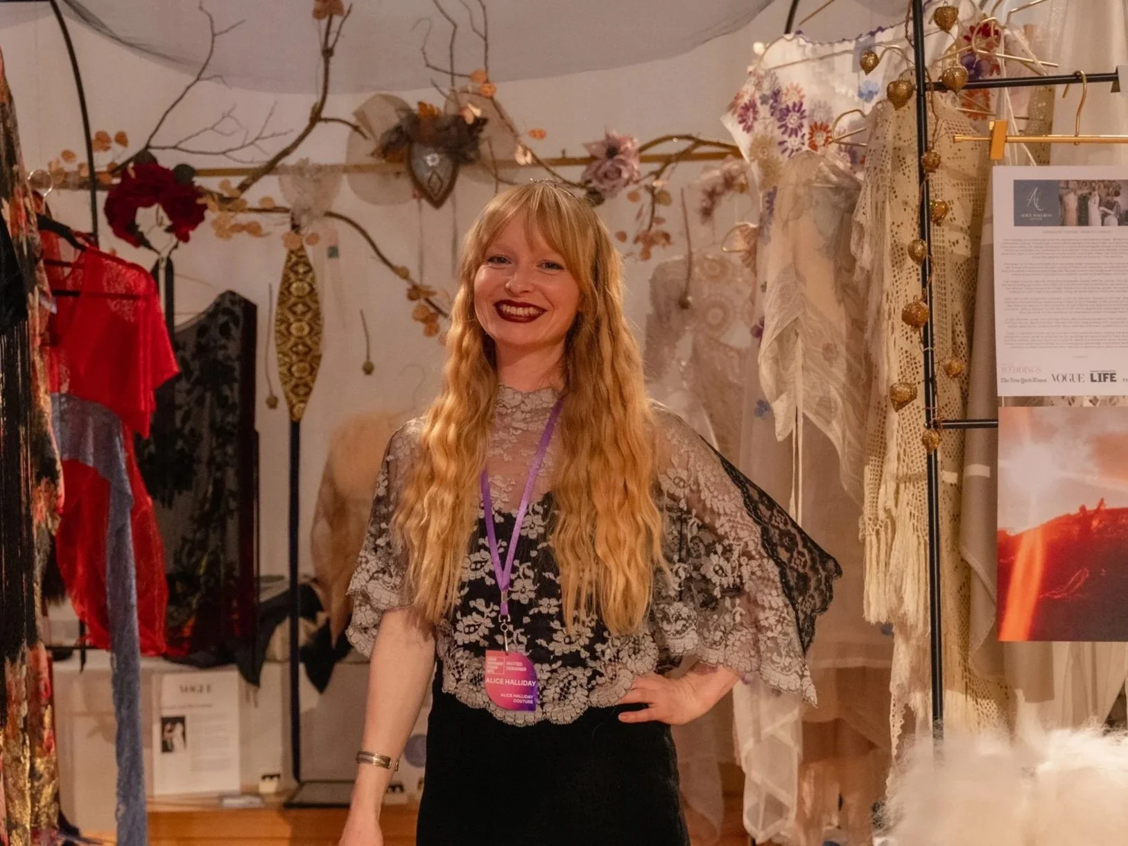 A smiling woman with long wavy red hair, wearing a lace top and a name badge, standing in a boutique or exhibition space surrounded by hanging fabric, lace, and decorative items.