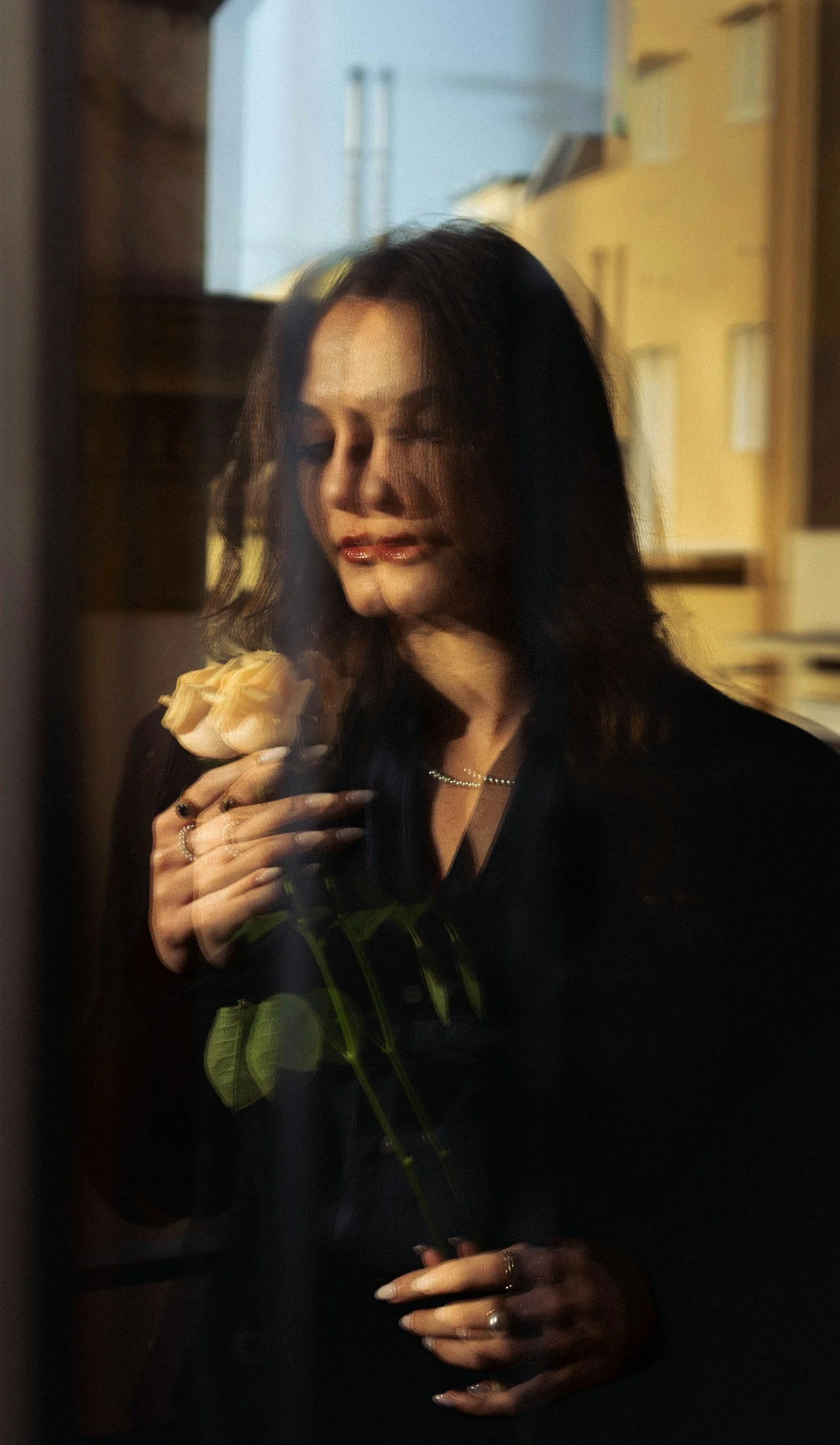 Woman with dark hair, closed eyes, holding a pale pink/orange rose, standing behind glass, with a blurred kitchen in the background.