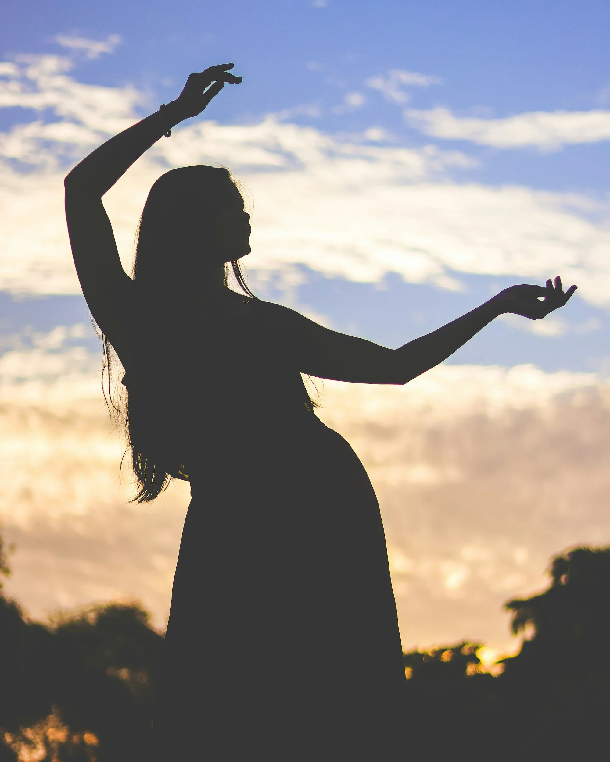 Silhouette of a pregnant woman standing outdoors during sunset, with arms raised and outstretched in a graceful pose, against a sky with clouds and trees in the background.
