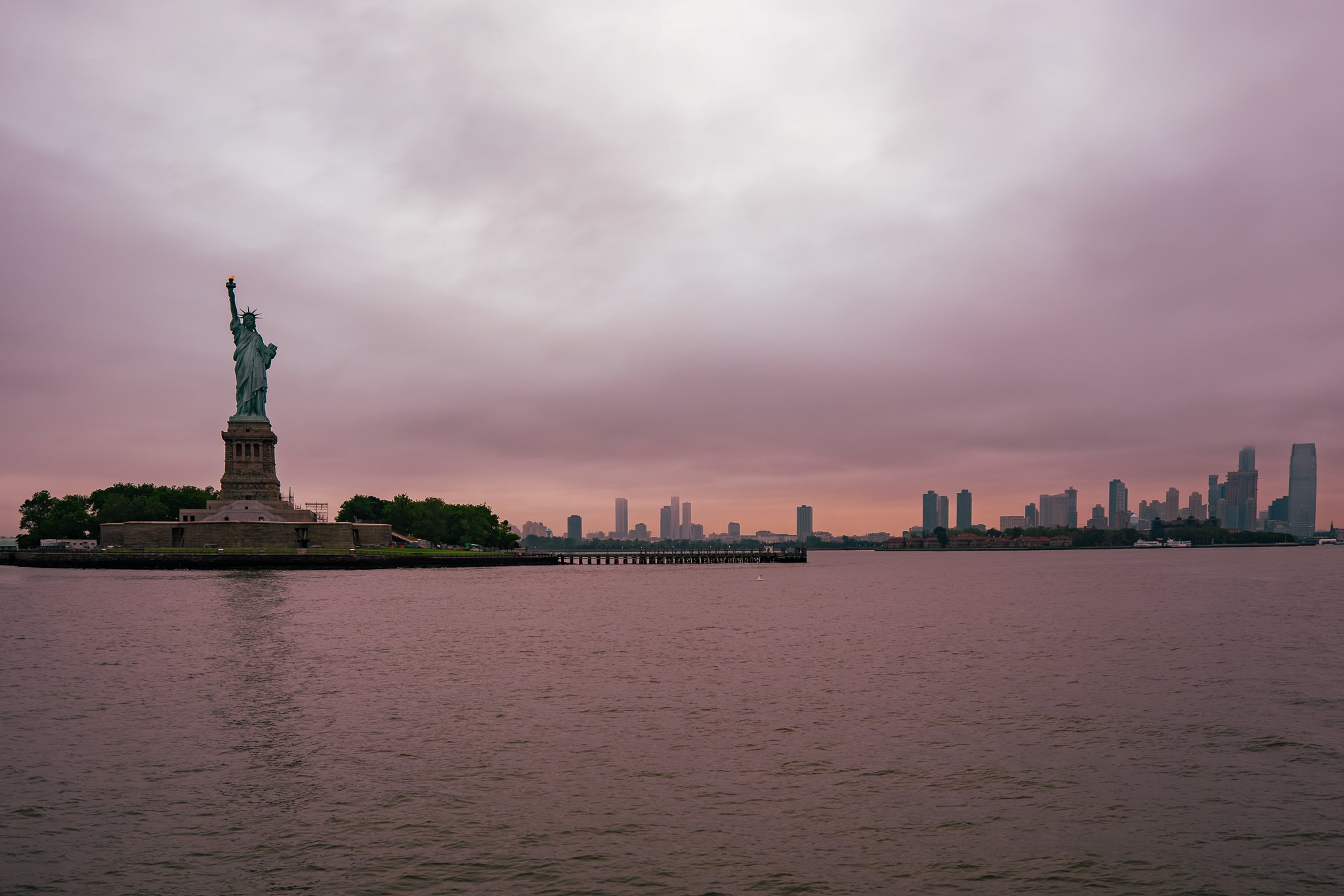 View of the Statue of Liberty on Liberty Island with New York City skyline and tall buildings in the background, under a cloudy sky.