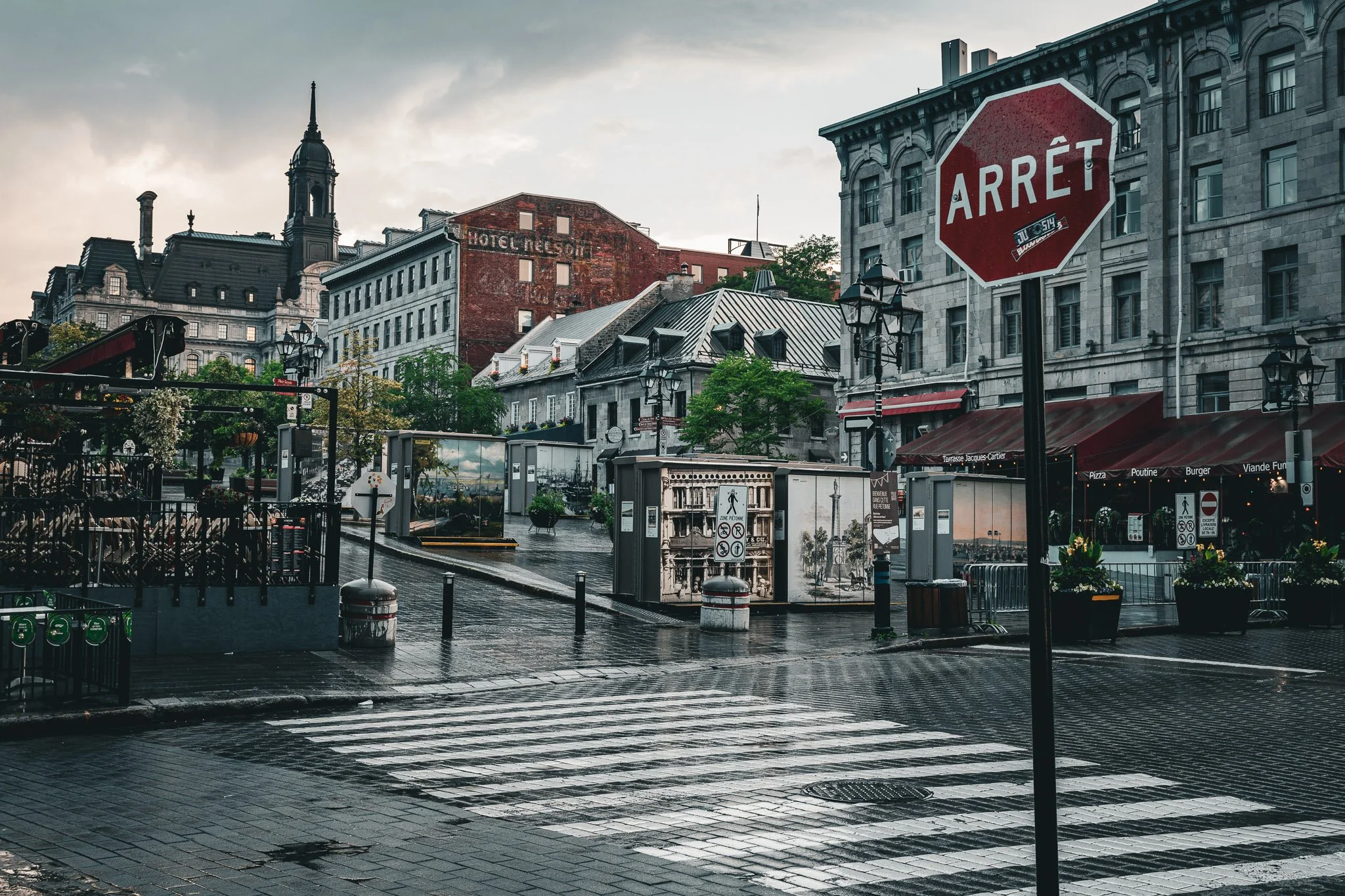 City street scene after rain with wet pavement, stop sign in French reading 'ARRÊT', historic European-style buildings, outdoor seating at a restaurant, and cloudy sky.