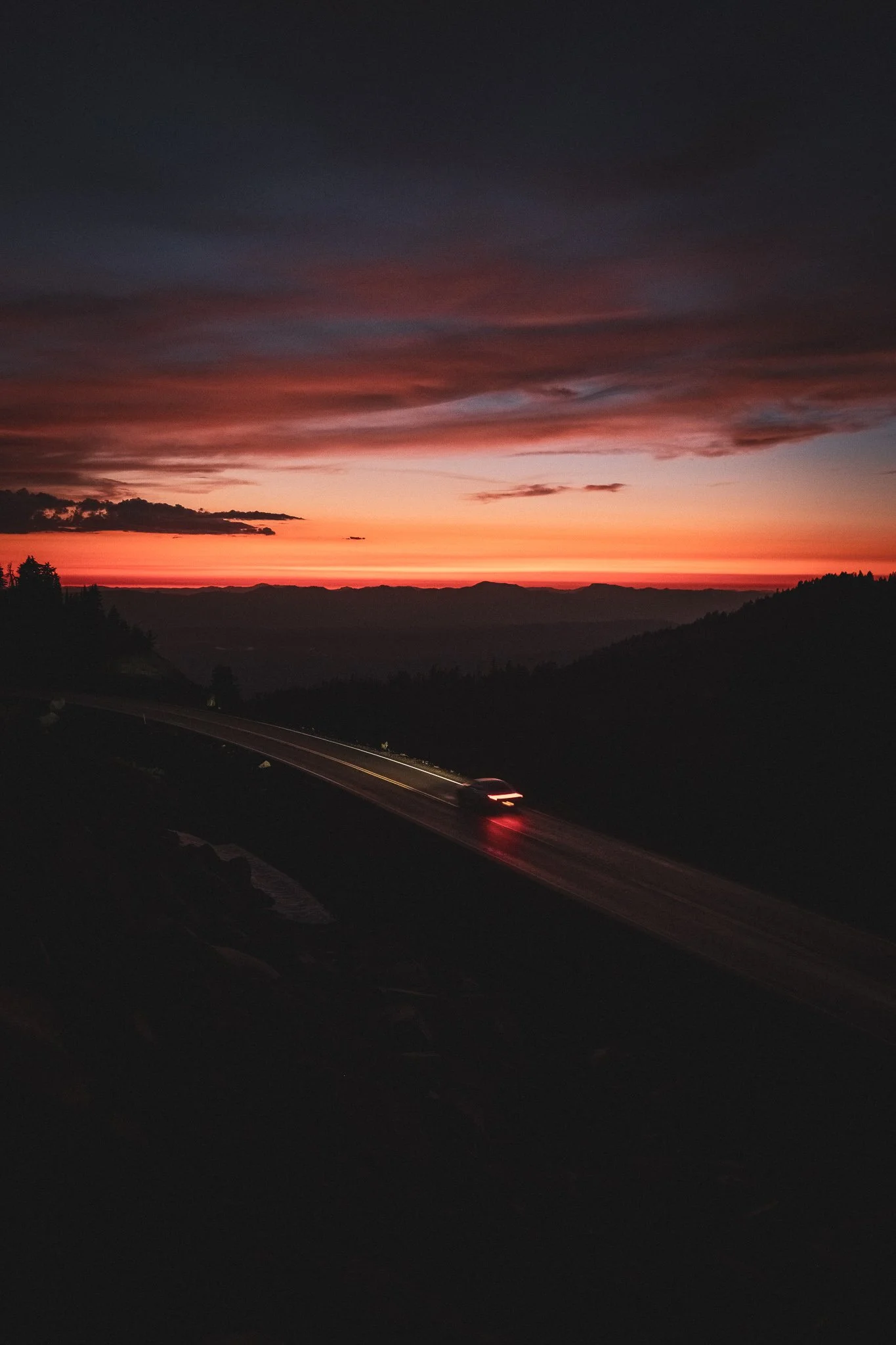A scenic view of a sunset over hills with a winding road in the foreground. A vehicle with headlights on is traveling on the road. The sky is filled with orange, pink, and dark clouds.