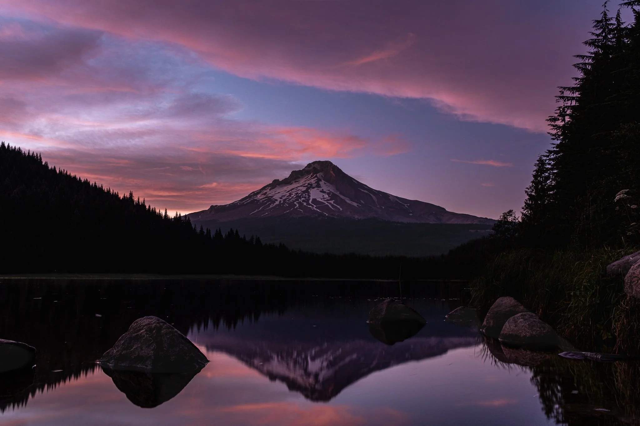 Mount Hood at sunset with colorful pink and purple clouds in the sky, reflected in a calm lake surrounded by rocks and pine trees.