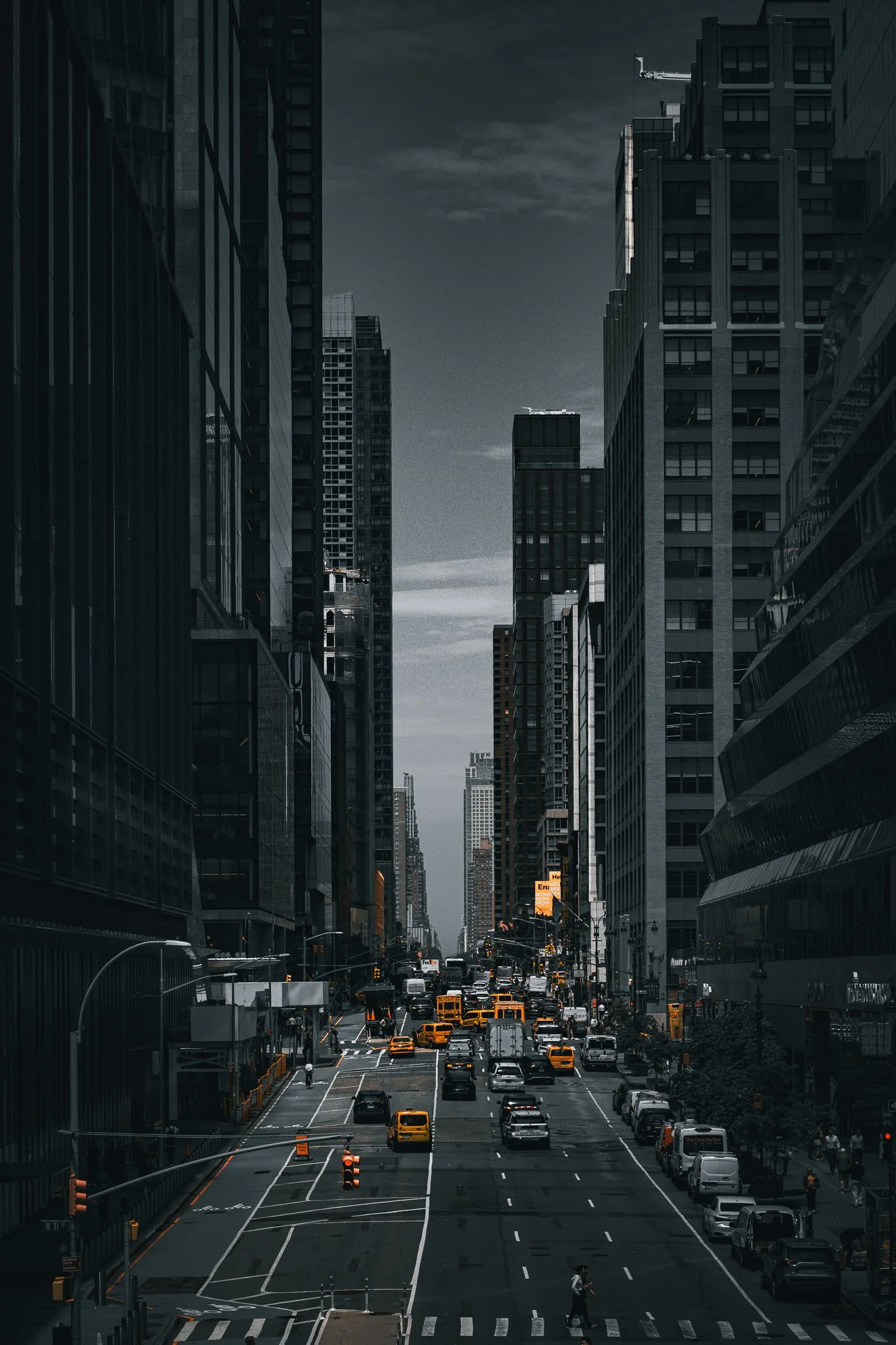 A city street with tall skyscrapers on both sides, yellow taxis, cars, and people walking, under a cloudy sky.
