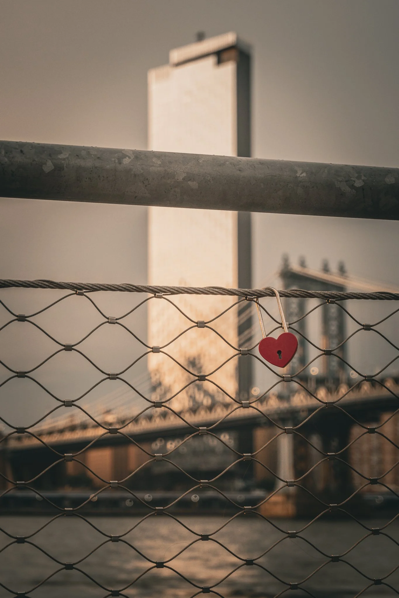 A close-up shot of a metal fence with a red heart-shaped padlock attached, with a city skyline and a bridge in the background during sunset.