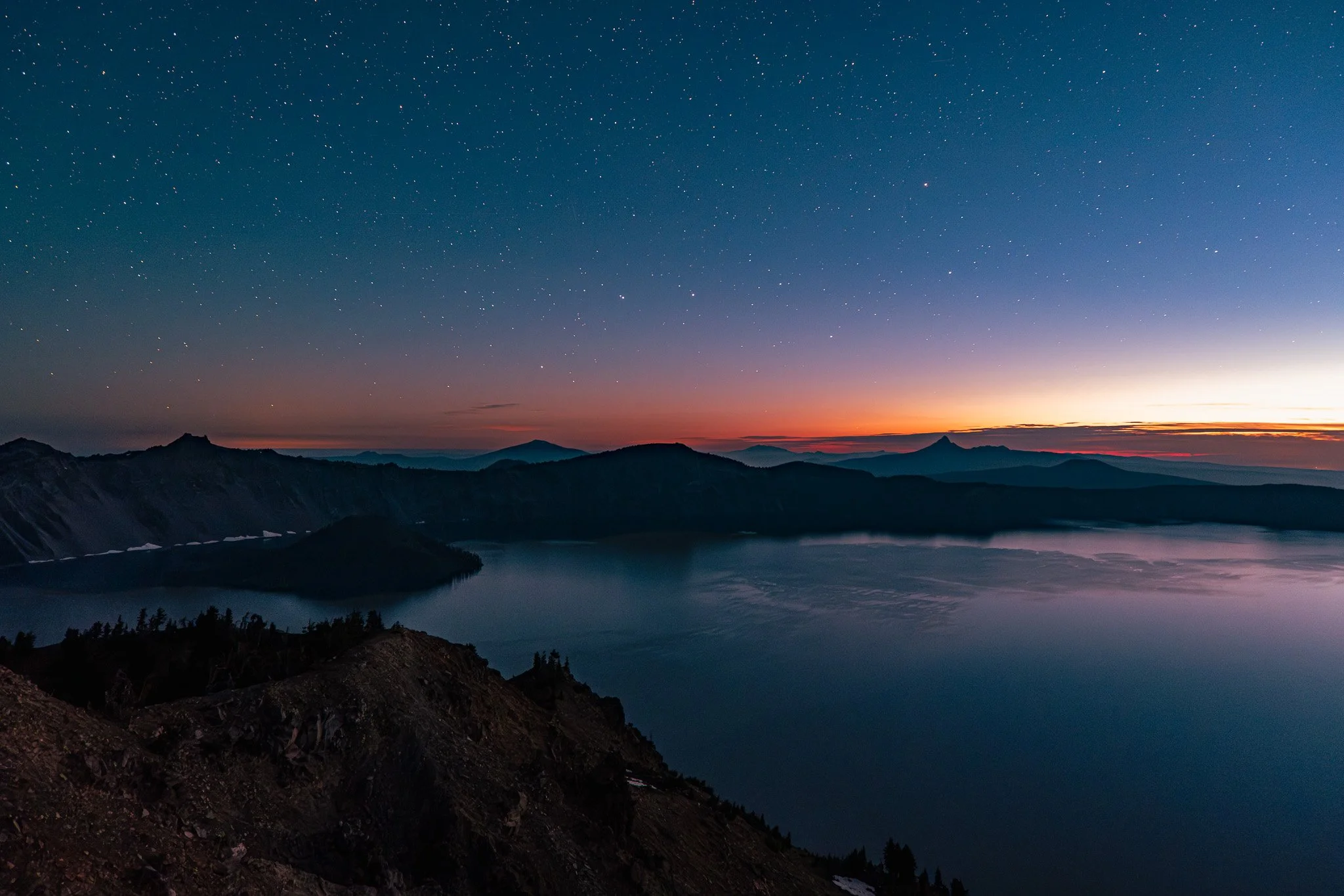 Nighttime view of a lake surrounded by mountains under a starry sky with a faint sunset glow on the horizon.