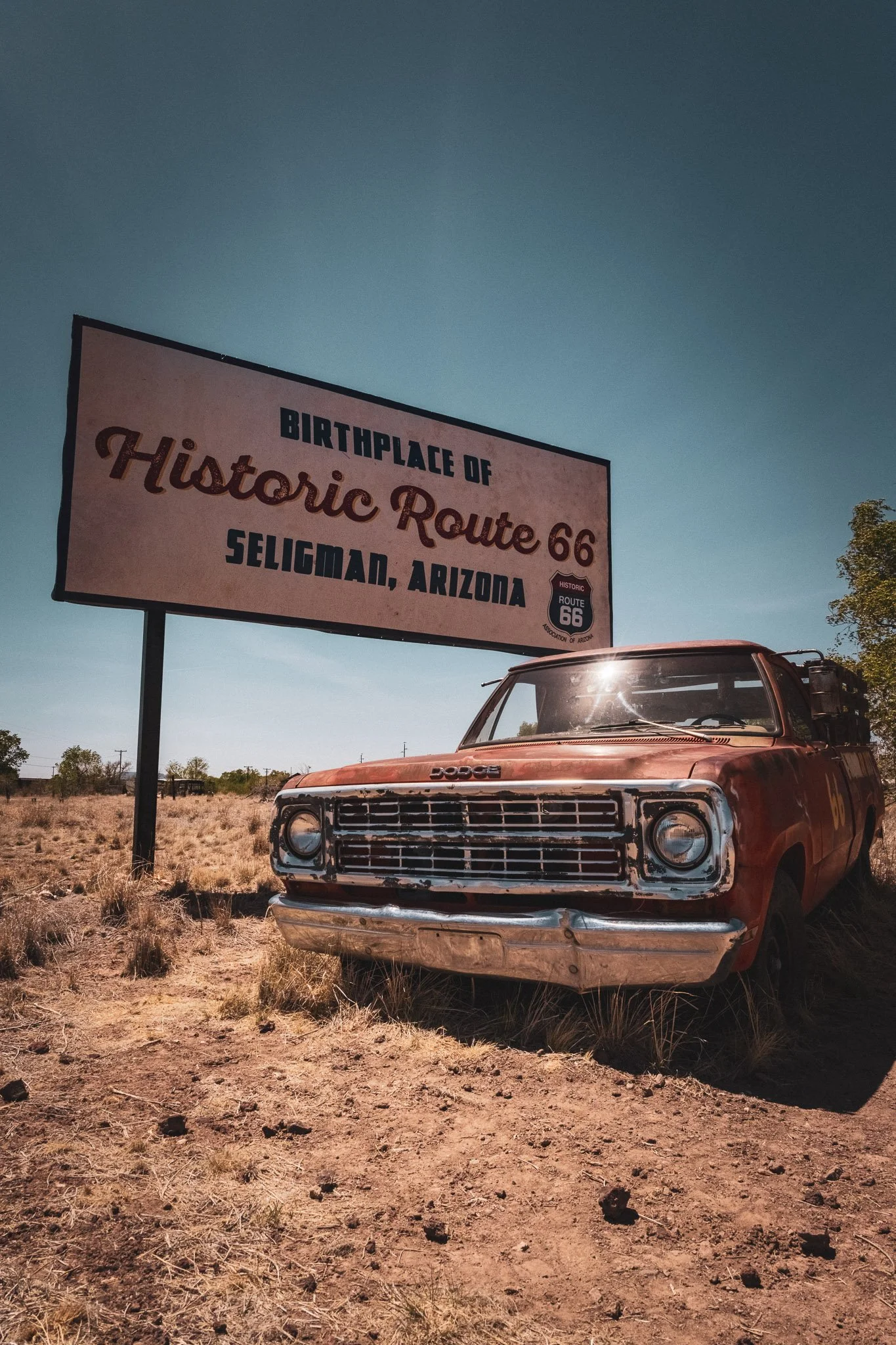 An old, rusty pickup truck parked on dry land next to a large sign that reads "Birthplace of Historic Route 66, Seligman, Arizona."