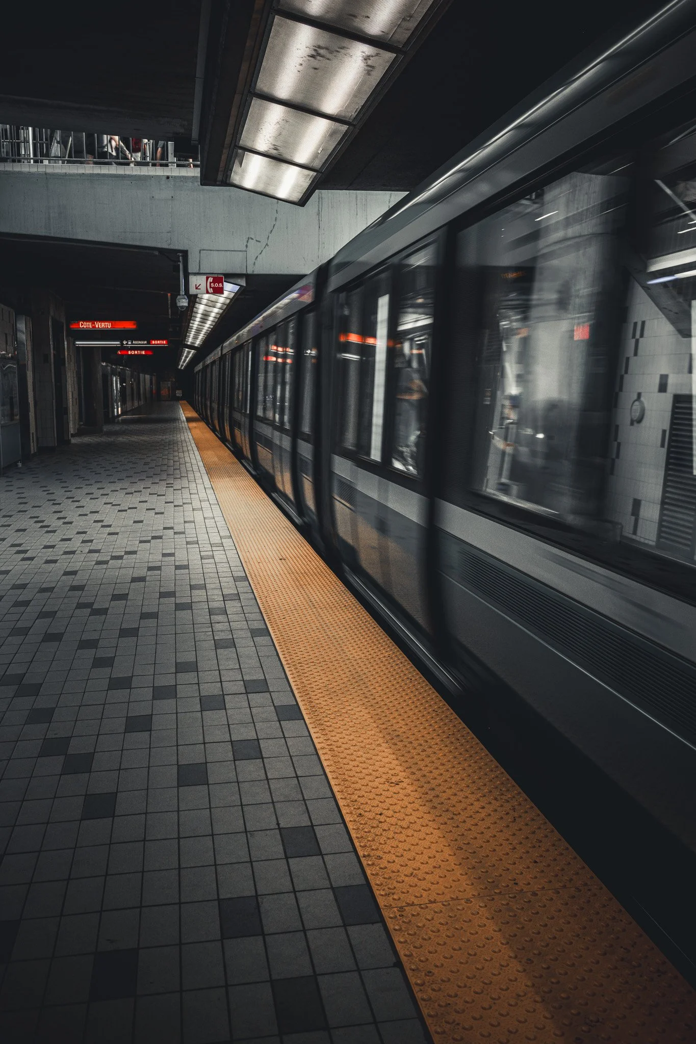 Subway train arriving at station with yellow tactile paving on platform, dark ceiling, and illuminated signs indicating direction and station name.