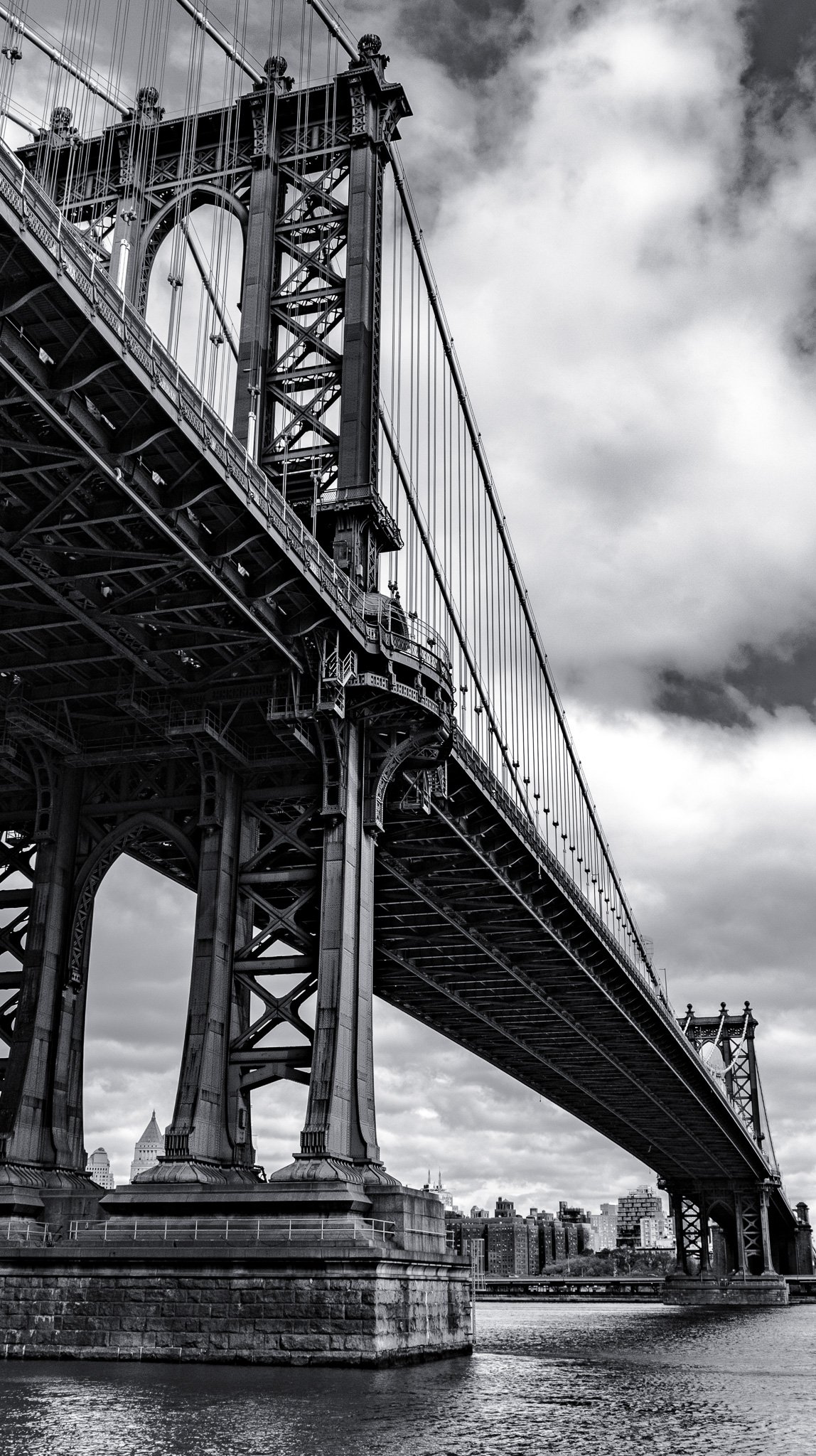 Black and white photo of the Brooklyn Bridge in New York City, showing its intricate steel structure and cables, with cloudy sky in the background and water below.