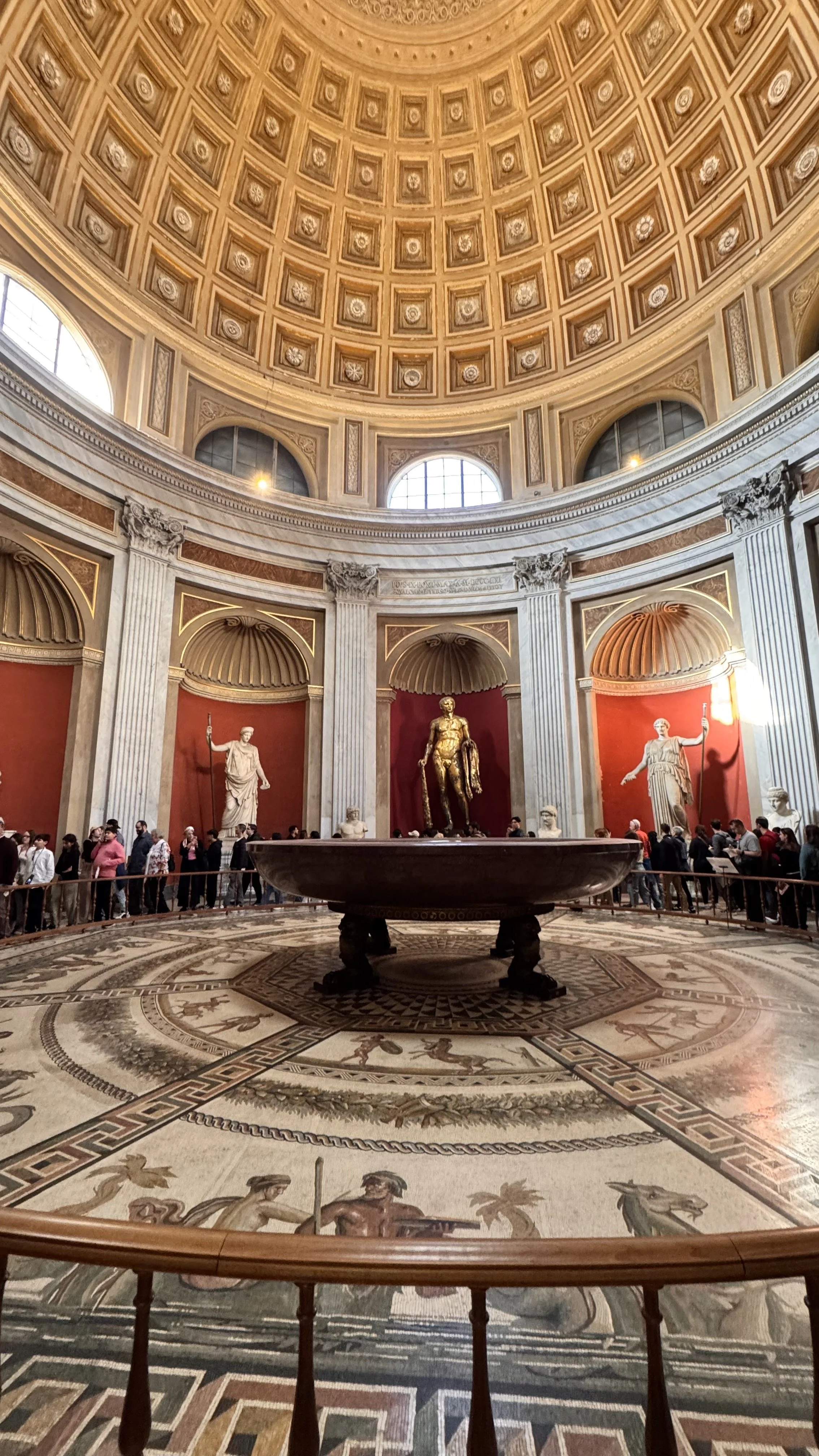 Interior of the Rotunda of the Vatican, featuring a domed ceiling, three statues behind red alcoves, a large fountain with a statue of a man in the center, and a patterned mosaic floor. Visitors are lined up along a barrier.