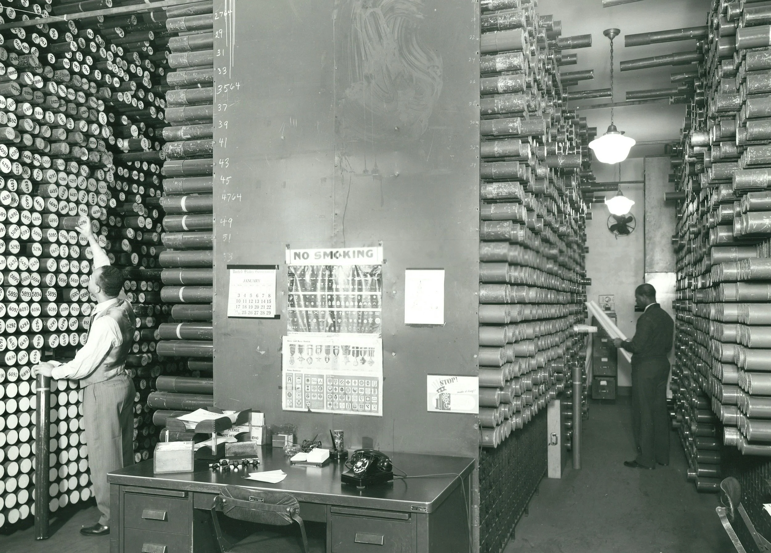 A black and white photo of a warehouse filled with tightly packed tall shelves of rolled-up papers or posters. Two men are seen, one on the left arranging items on a shelf and the other on the right reading a large document. There are office items on a desk, signs on the wall including a 'NO SMOKING' sign, and hanging ceiling lights illuminating the space.