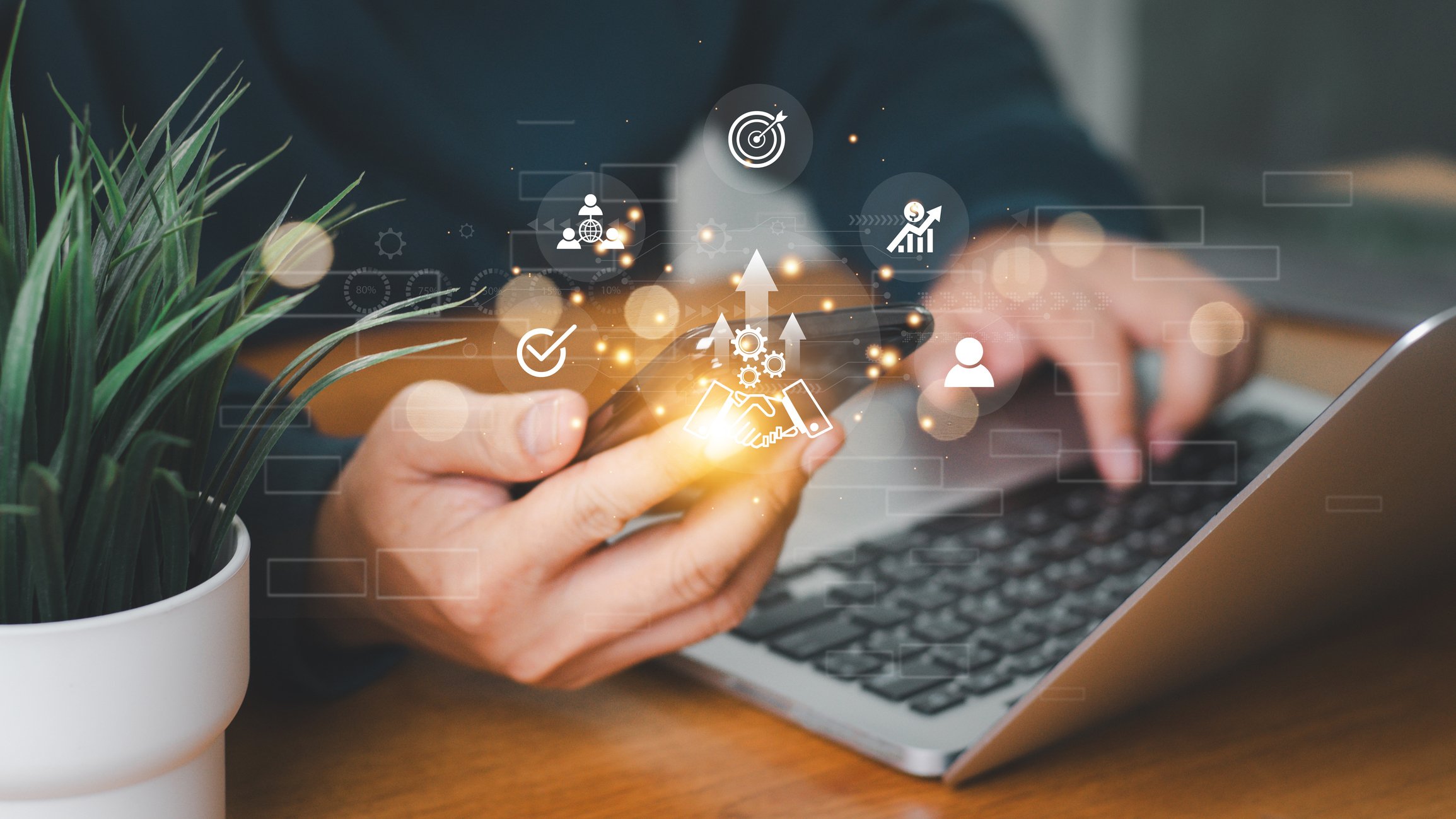 Person using smartphone with digital icons and graphs overlay, workspace includes a laptop and a potted plant.