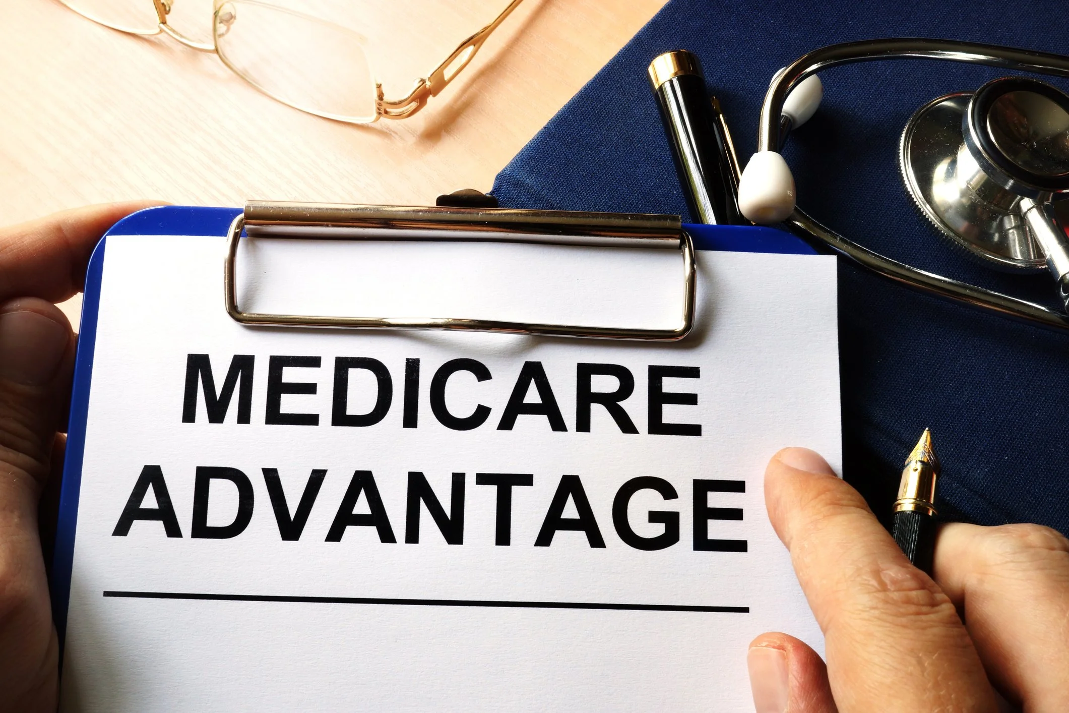 A person holding a clipboard with the words "MEDICARE ADVANTAGE" on it, next to a stethoscope and a pen, with eyeglasses and a blue cloth in the background.