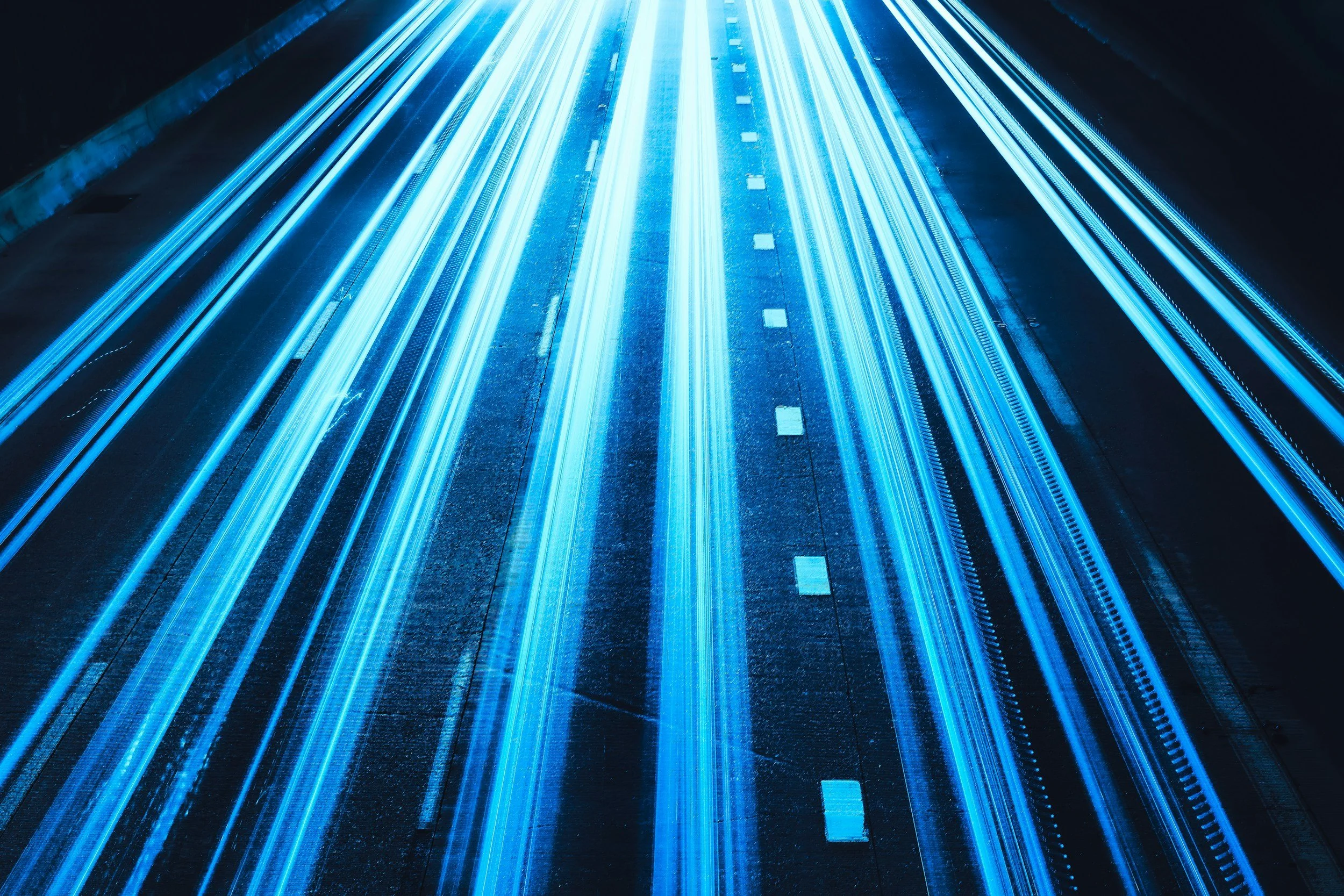 Long exposure photograph of a highway at night with streaks of bright blue and white lights from moving vehicles.