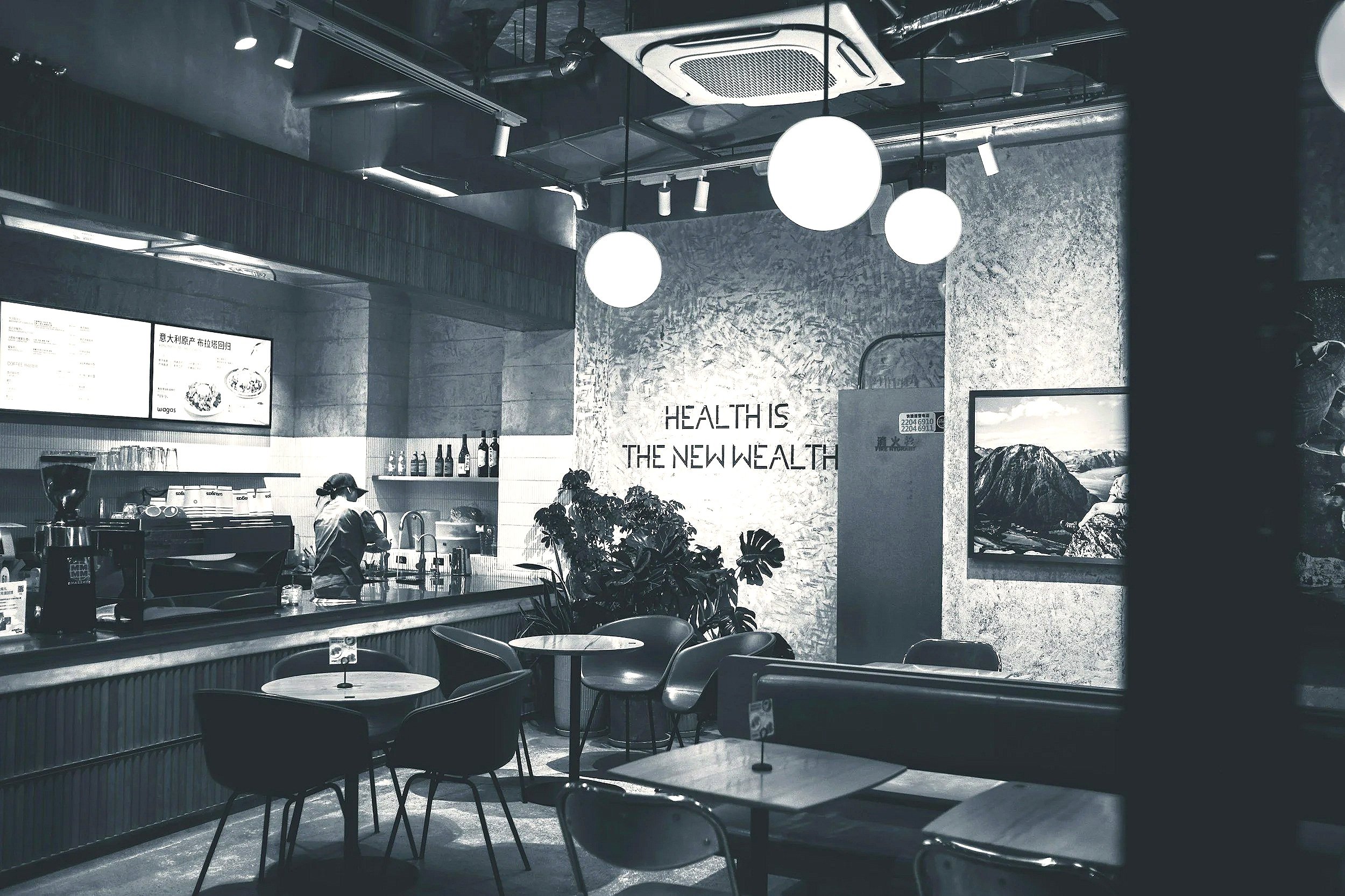 Black and white photo of a modern cafe or coffee shop interior with tables, chairs, a barista working behind the counter, and a wall with the phrase 'Health is the new wealth' written on it.