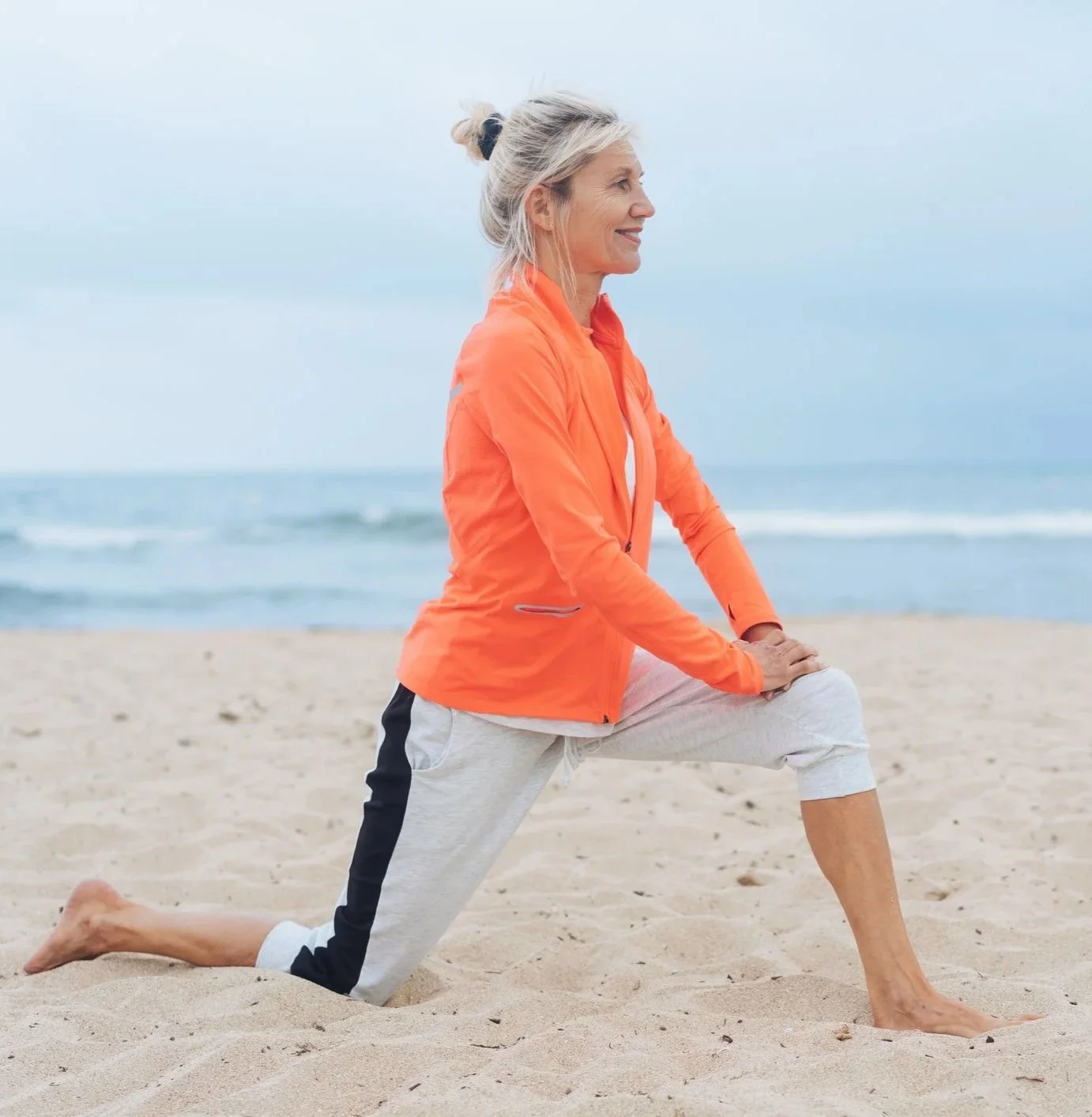 An older woman practicing yoga on a sandy beach, wearing an orange jacket and gray pants, with the ocean and cloudy sky in the background.