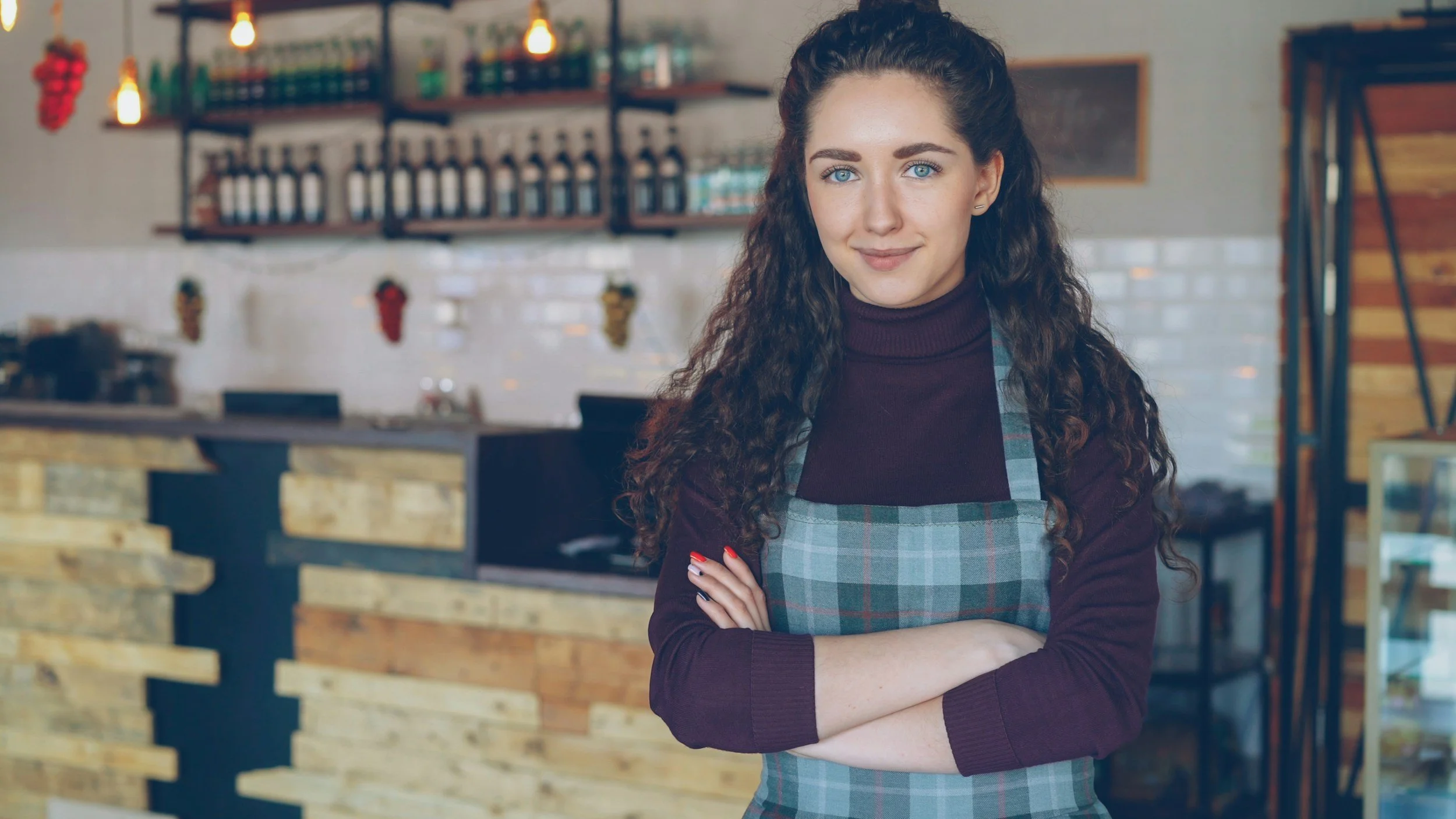 A woman with long curly brown hair and blue eyes standing inside a cafe, wearing a plaid apron over a maroon turtleneck, smiling with arms crossed.
