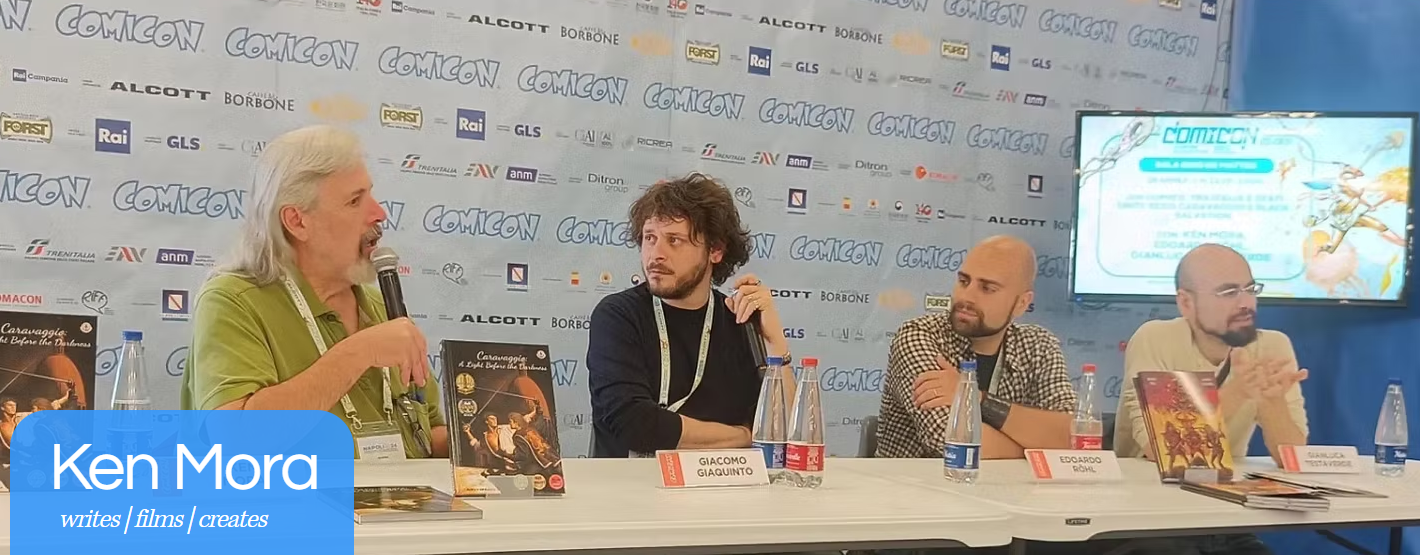 Four men sitting at a panel table during a conference. The man on the far left, with long gray hair, is speaking into a microphone. Each has a nameplate and hopoge, graphic novel, or book in front of them. A background banner displays the "COMICON" logo repeatedly. There is a large screen to the right showing a colorful animated graphic with indistinct text.