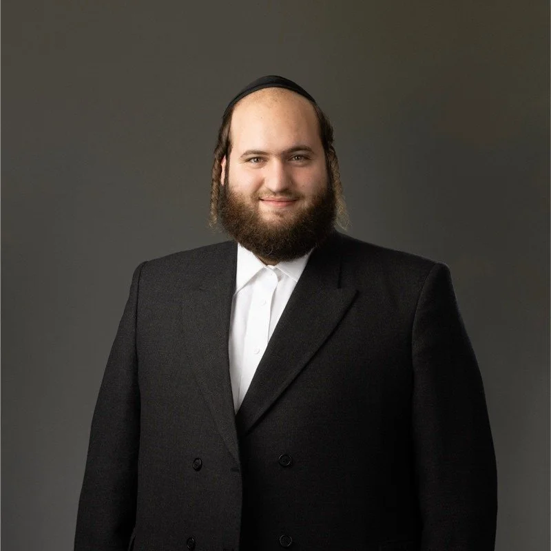 A man with a beard and long hair, wearing a black suit and a white shirt, smiling at the camera against a plain gray background.