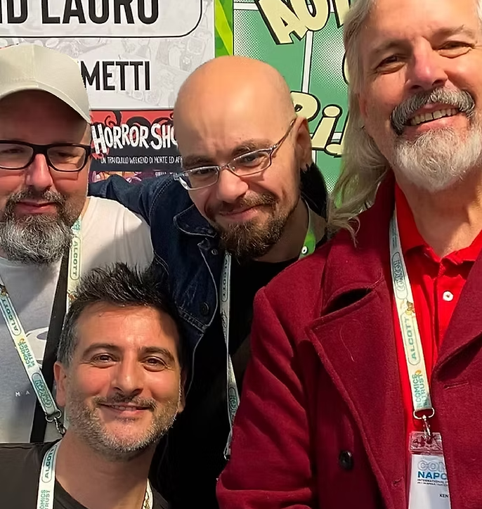 Four men smiling for a selfie at a convention, with various posters and signs in the background.