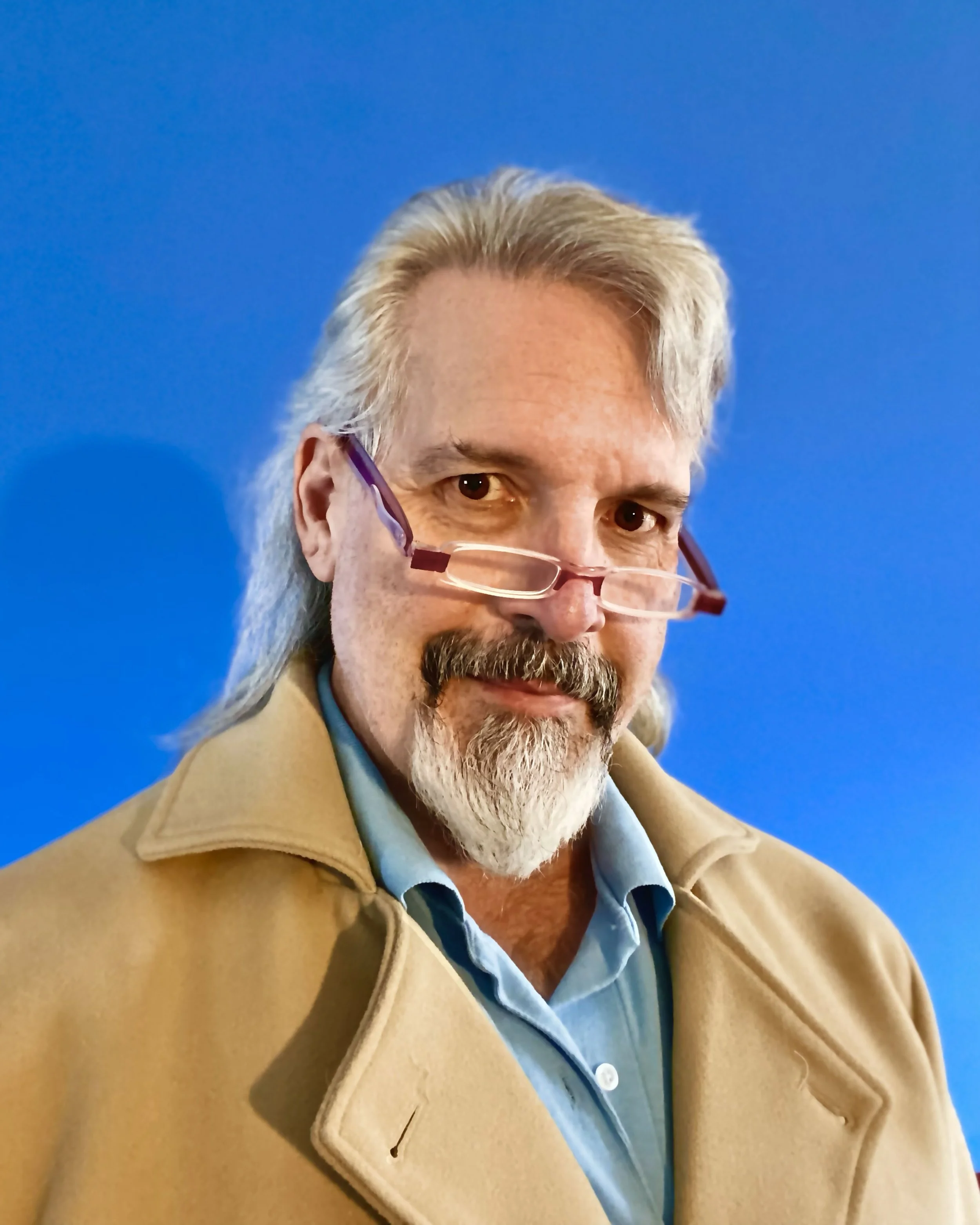 A man with gray hair and a beard wearing glasses, a tan coat, and a light blue shirt standing against a blue background.
