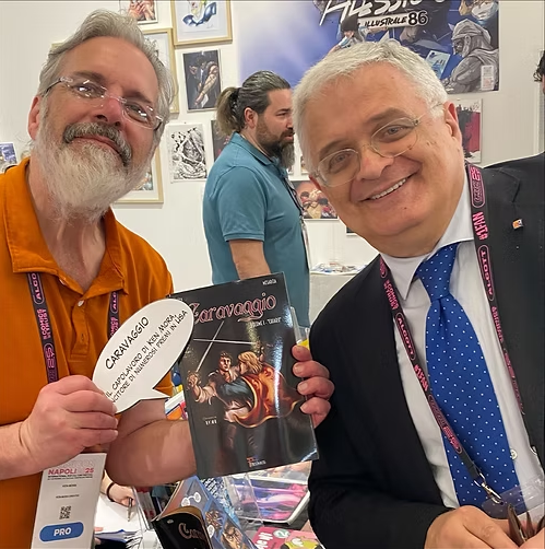 Two men smiling and holding a comic book and a speech bubble at a comic convention. The man on the left has a beard and glasses, wearing an orange shirt. The man on the right has gray hair, glasses, and is dressed in a suit with a blue tie. A person 