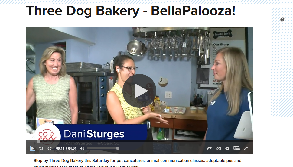 Three women are inside a bakery kitchen with stainless steel equipment and hanging utensils, engaged in a conversation. One woman is holding a microphone, and all three are smiling.