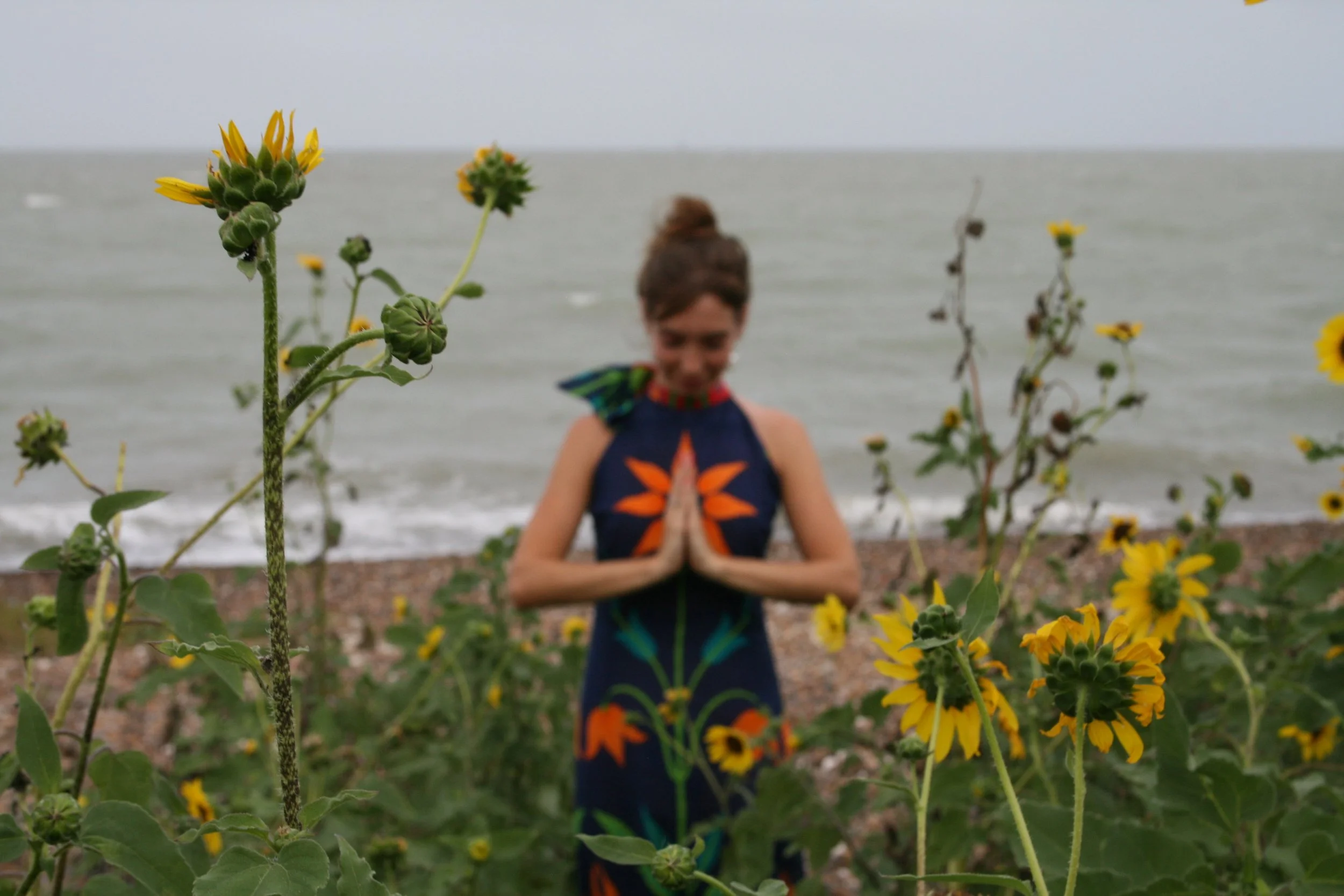 A woman practicing yoga or meditation on a beach, surrounded by yellow flowers, with the ocean in the background.