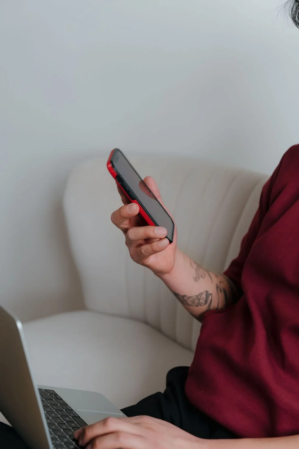 Person using a smartphone with a tattooed arm, seated on a light-colored sofa, with a laptop on their lap.
