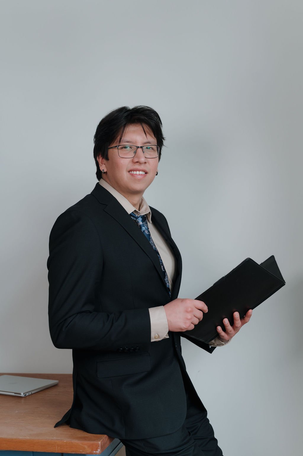A young man with dark hair, glasses, wearing a black suit, beige shirt, and a patterned tie, holding a black folder, standing next to a wooden desk with a closed laptop on it against a plain wall.