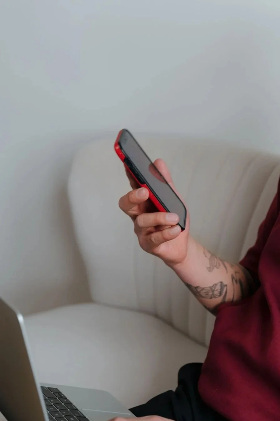 Person holding a smartphone with a red case while sitting on a white chair next to a silver laptop.