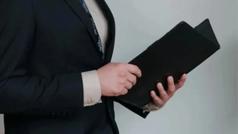 Close-up of a person in a dark suit holding a black folder or tablet, with the focus on their torso and hands.