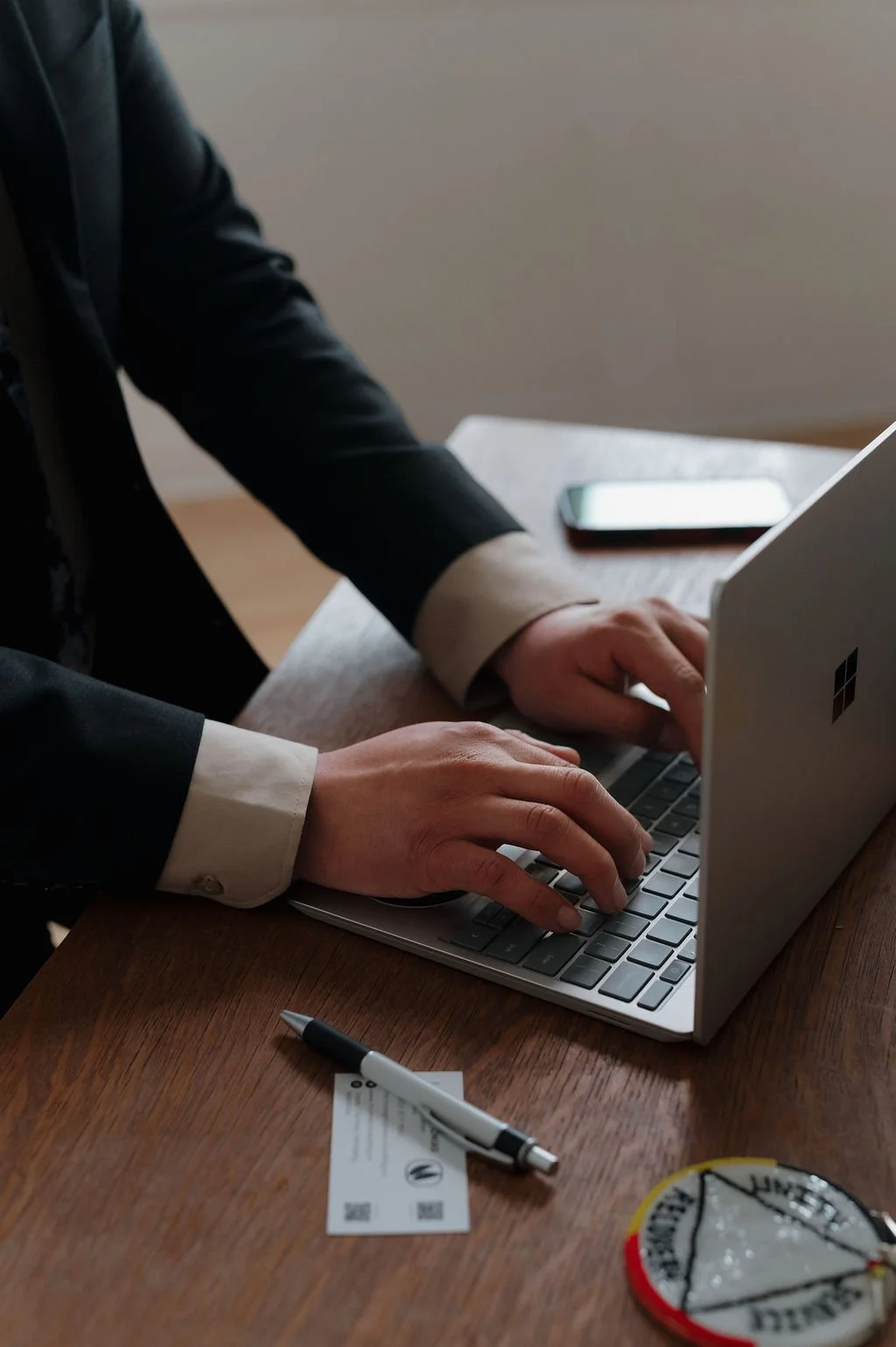 Person working on a Microsoft Surface Laptop at a wooden desk, with a business card, pen, a mobile phone, and a round patch or badge nearby.