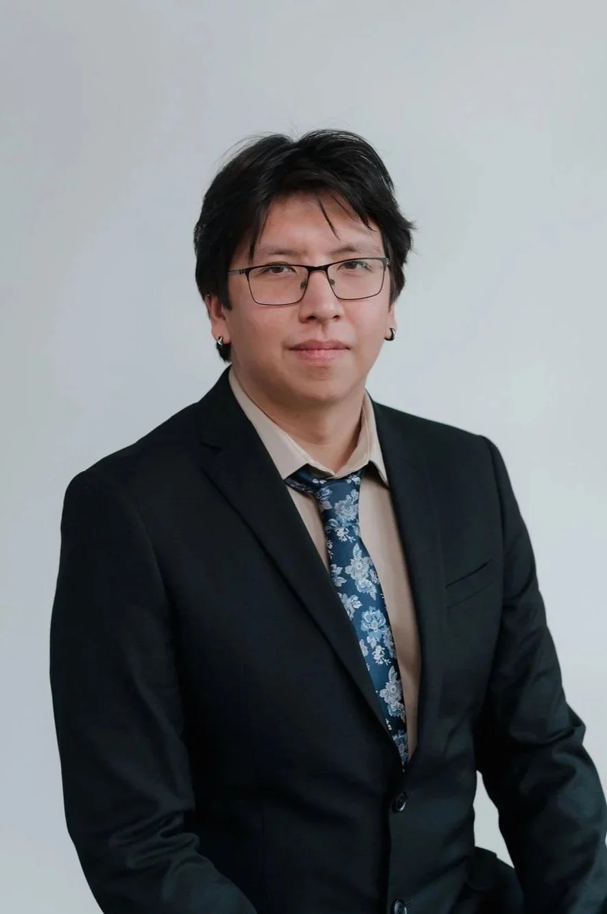 A young man with black hair, glasses, and earrings wearing a black suit, beige shirt, and blue floral tie posing against a plain light-colored background.