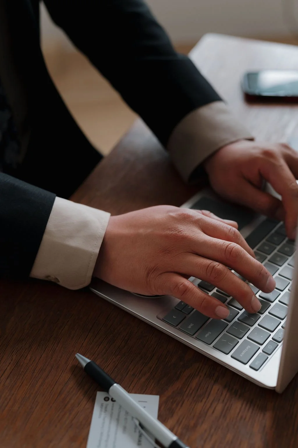 Person typing on a silver laptop at a wooden desk, with a pen and a smartphone nearby.