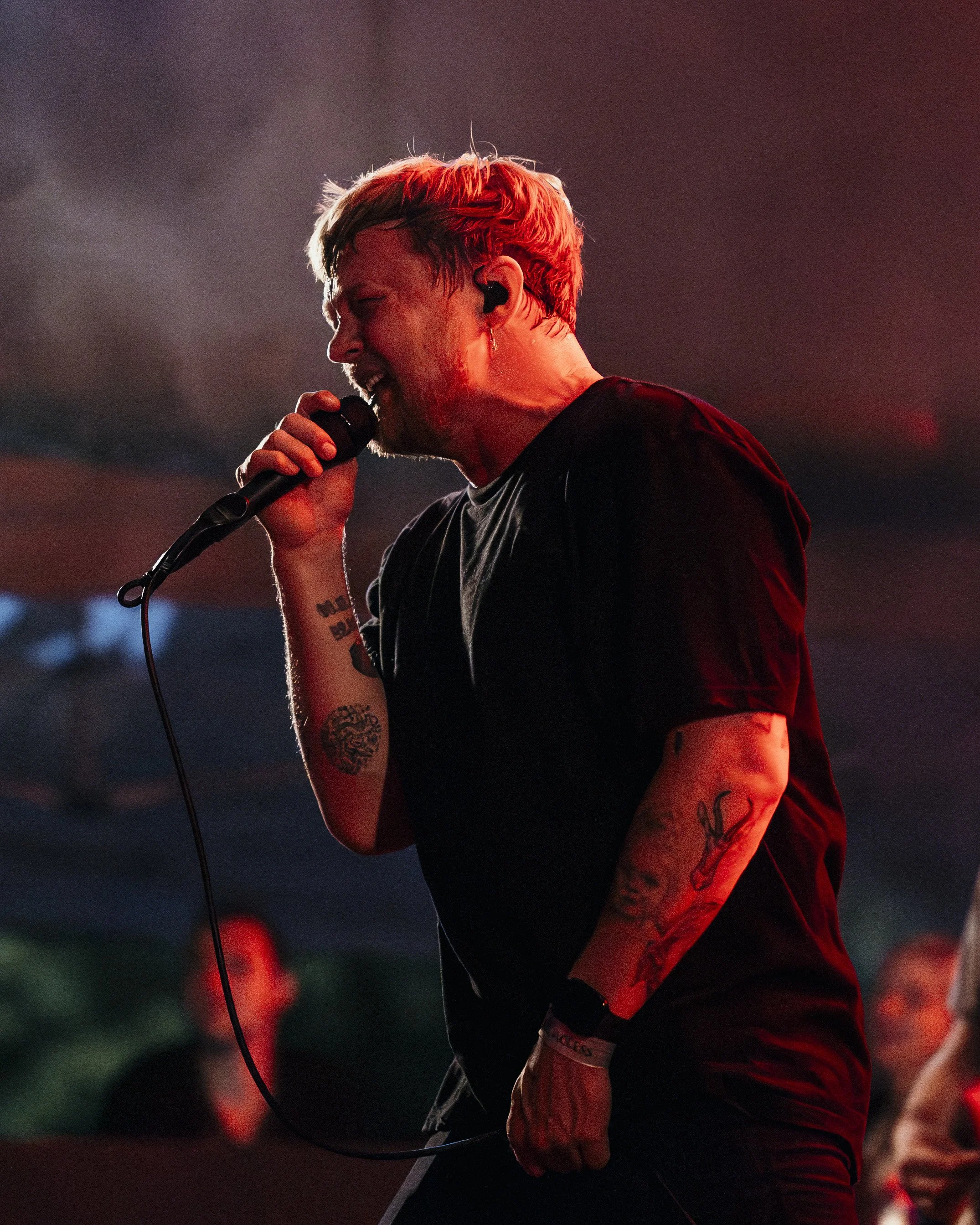 A man performing with a microphone on stage, with tattoos on his arms, wearing a black t-shirt, and red lighting overhead.