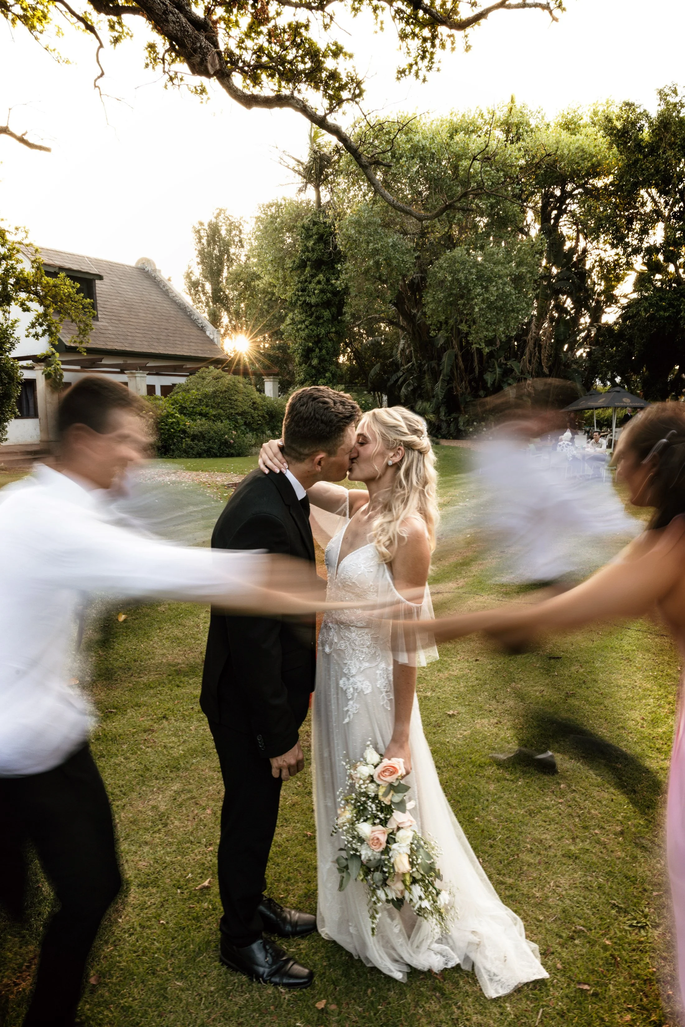 A bride and groom kiss outdoors during their wedding ceremony at sunset, while blurred wedding guests move around them.