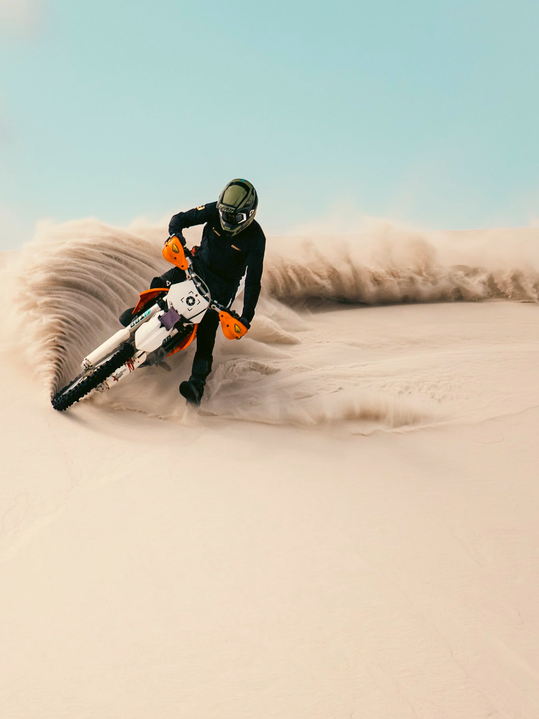 Person riding a dirt bike on a sandy, desert-like terrain with sand swirling around, wearing a helmet and black protective gear.