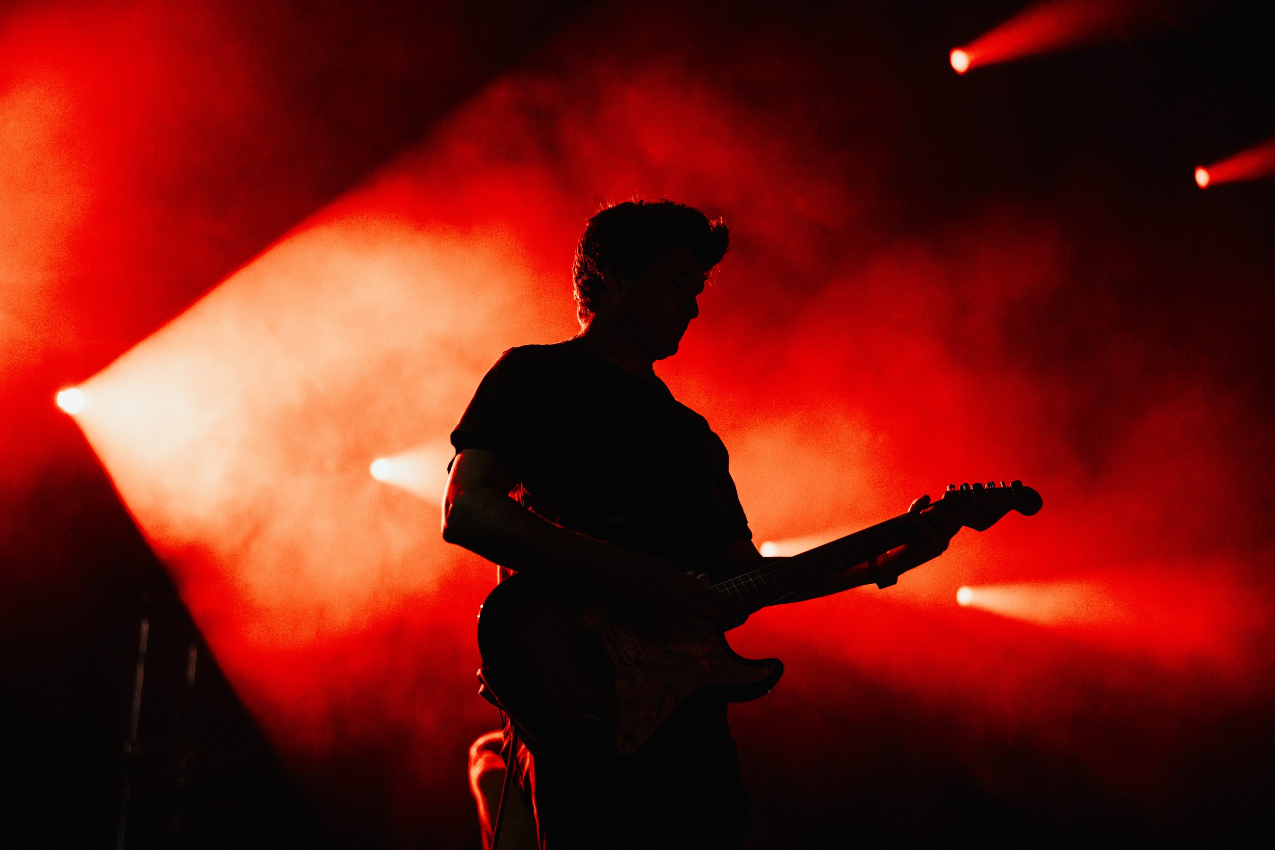 Silhouette of a person playing electric guitar on stage with red stage lighting and background smoke.