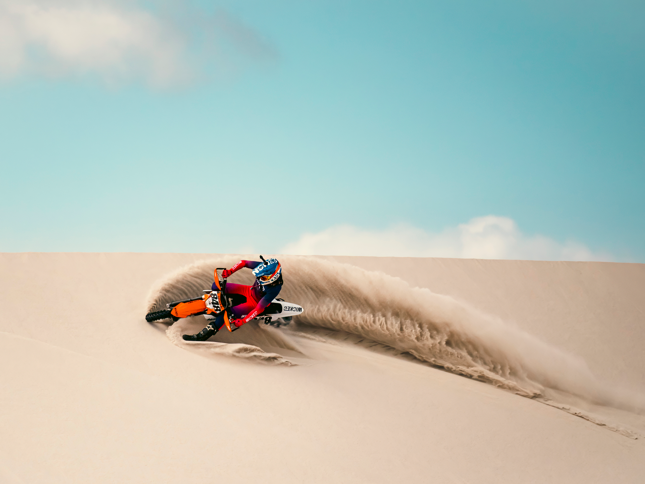 Motocross rider in red gear racing down a sand dune with a spray of sand behind him.