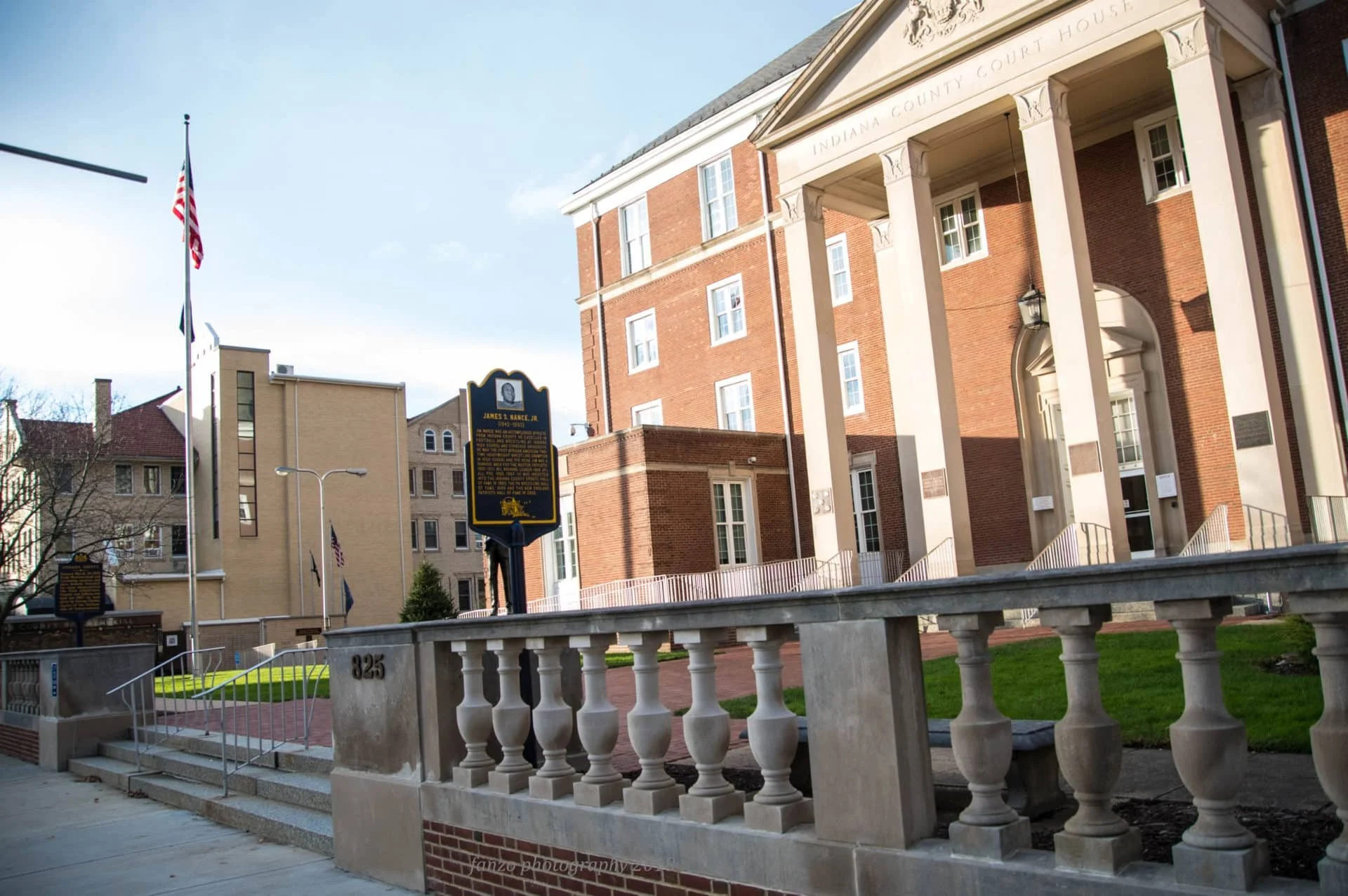 A historic brick courthouse with white columns in Indiana, with a black historical marker and American flags in front, and neighboring buildings, under a bright sky.