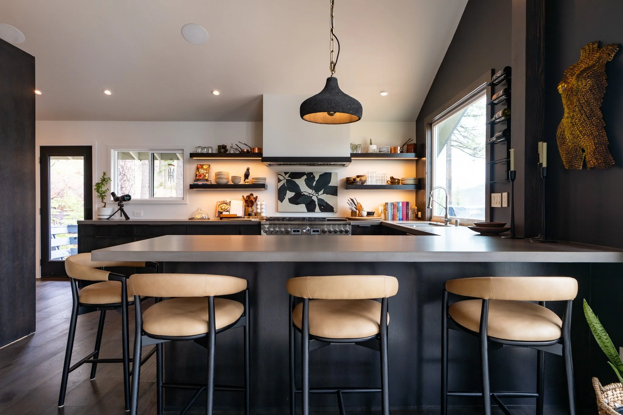 Modern kitchen with black and white decor, pendant light, open shelves with dishes, large window, and beige chairs around an island.